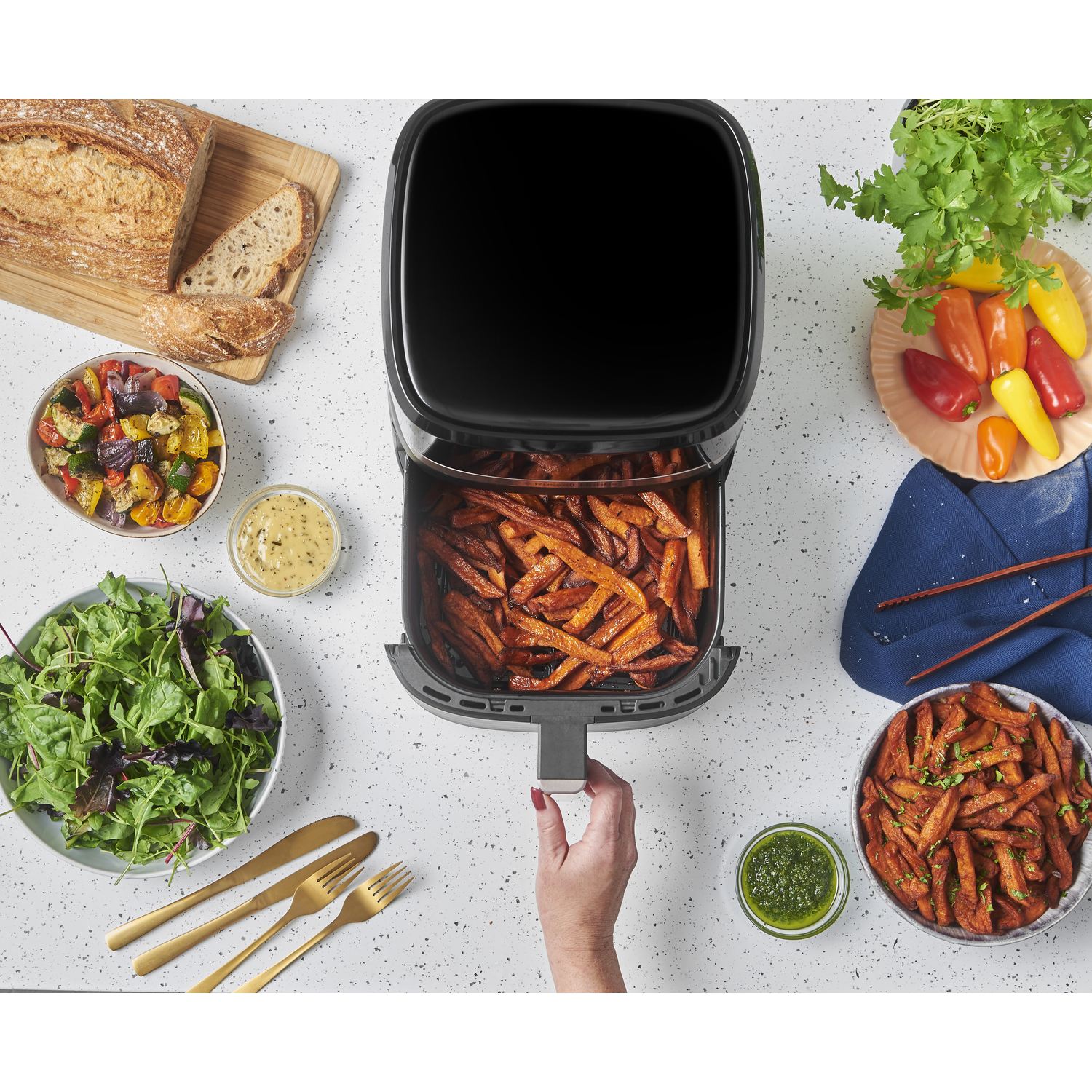 An air fryer with sweet potato fries, bread, salad, vegetables, and sauces on a white surface. A hand pulls out the fryer tray.
