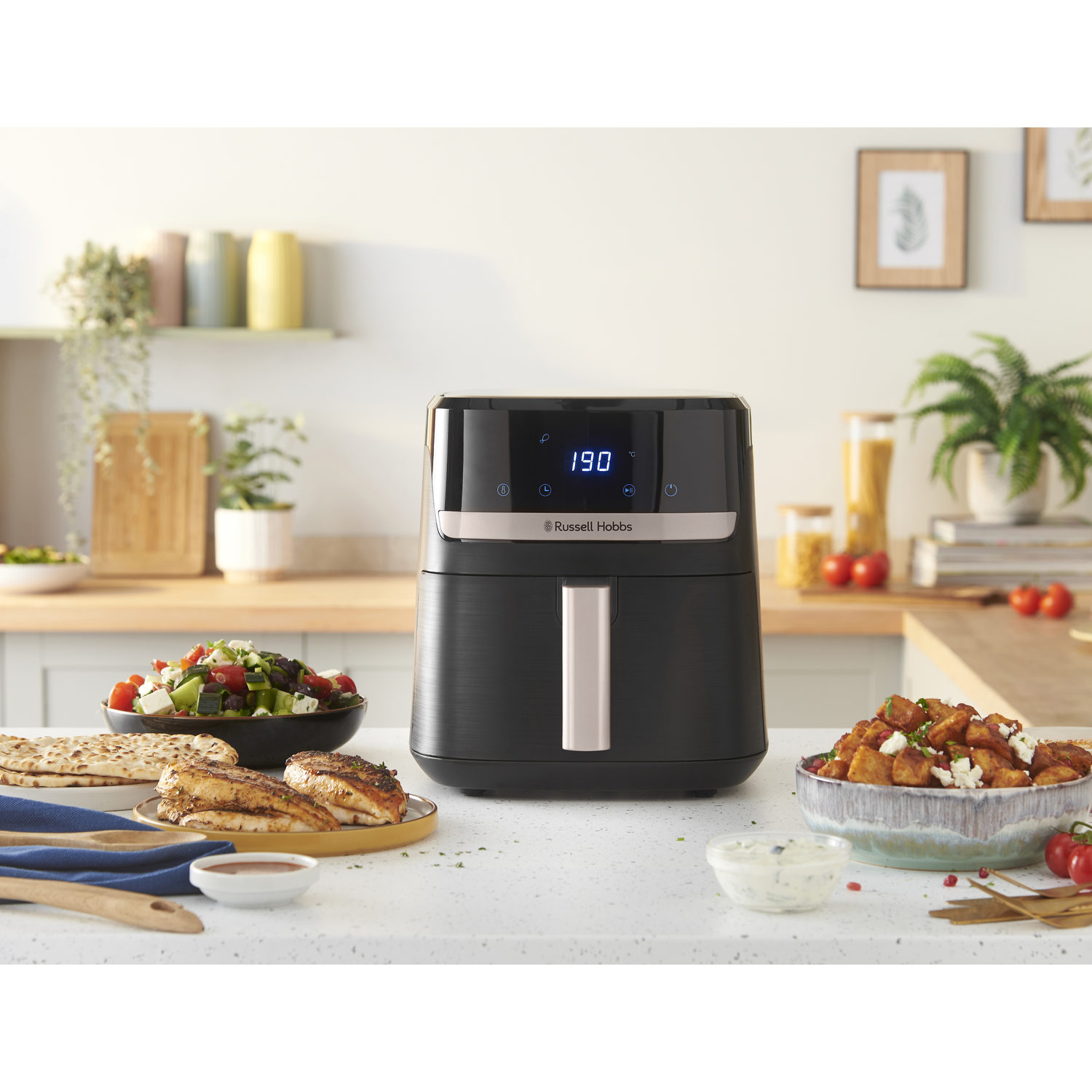 An air fryer with food on a table. Chicken, salad, and potatoes are visible. Background includes a kitchen setting.