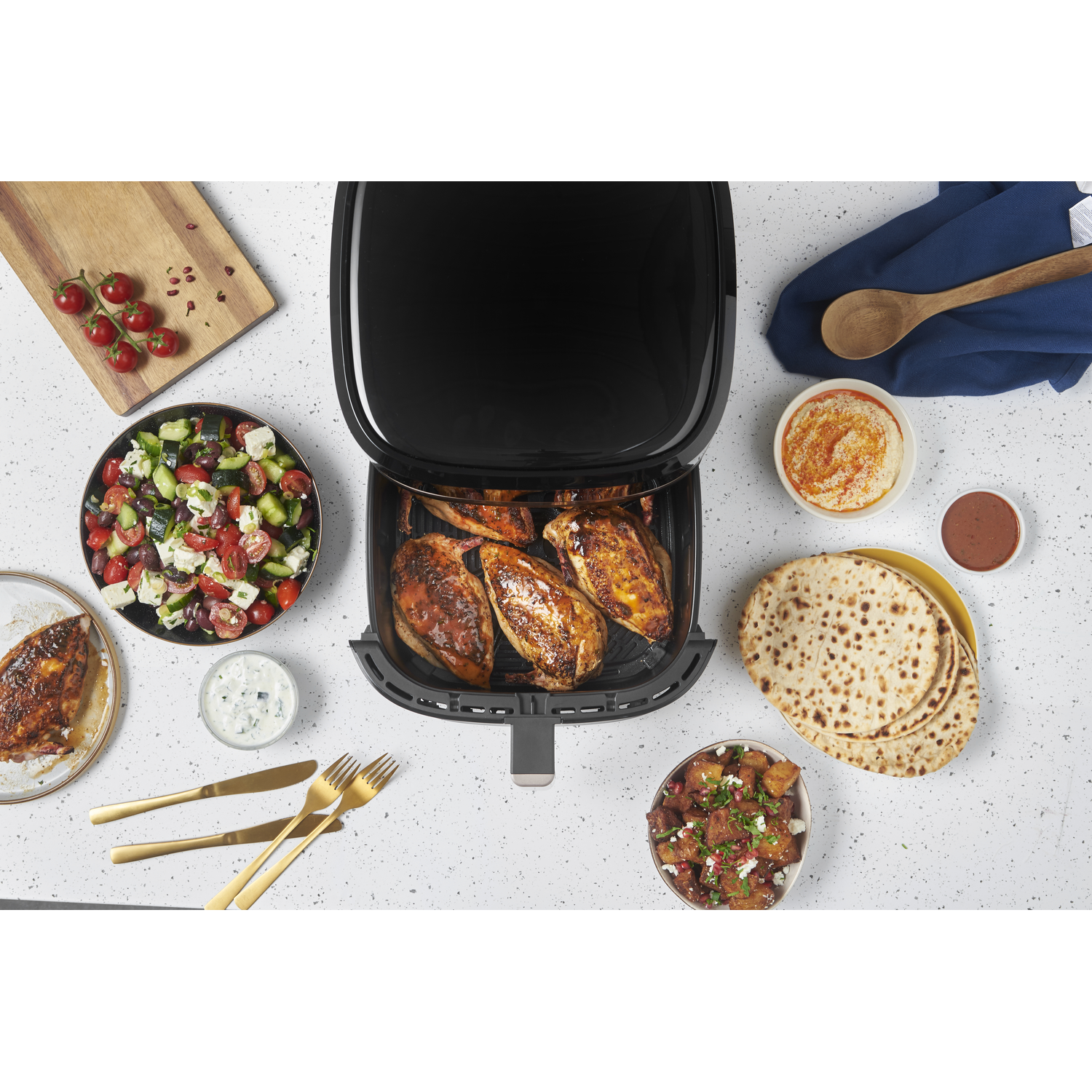 An air fryer with cooked chicken, surrounded by a salad, hummus, bread, potatoes, and tomatoes. Golden cutlery is on the left.