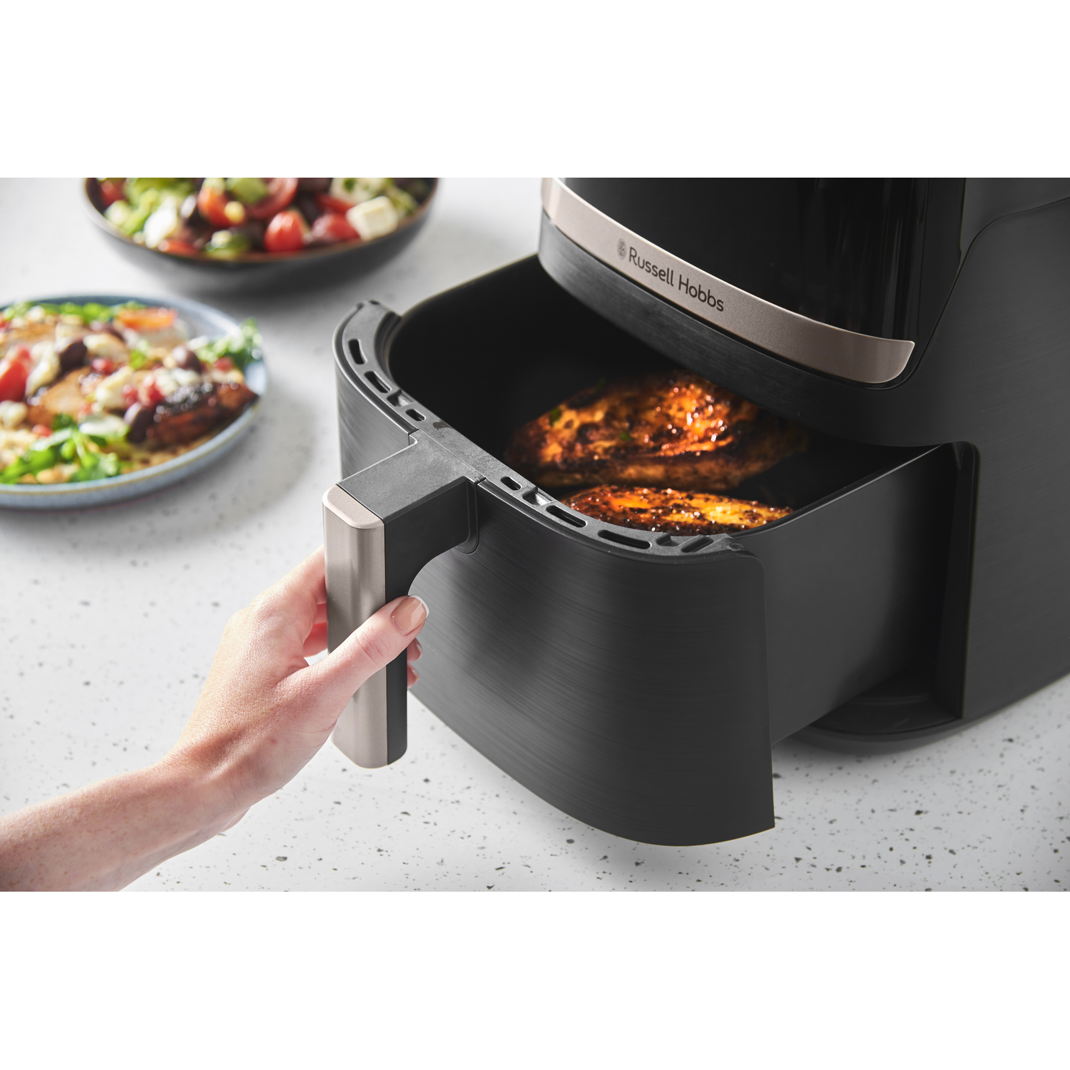 A hand pulls out an air fryer basket with cooked chicken. Salad bowls sit in the background.