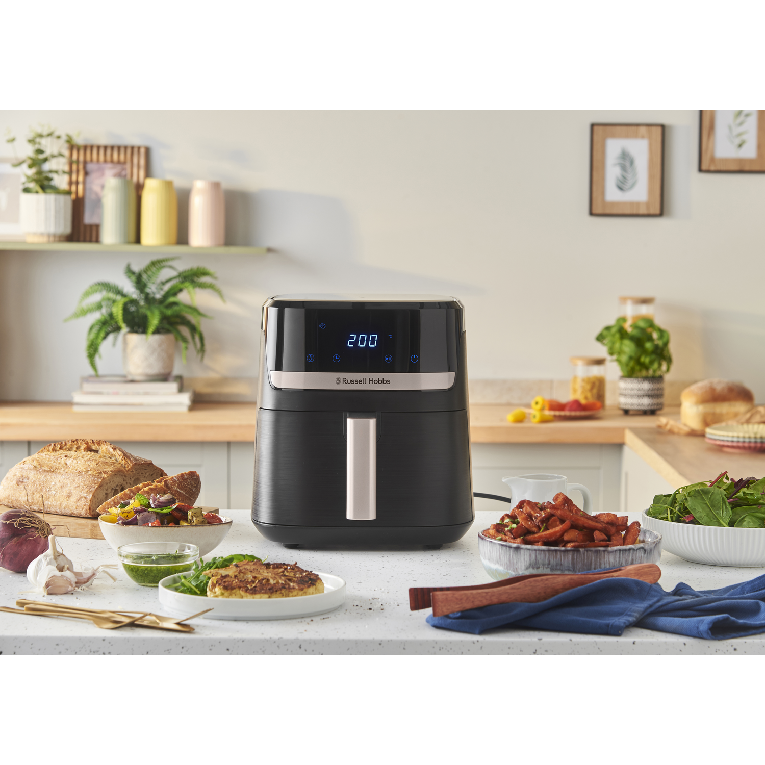 Black air fryer with food on a counter. Bread, salad, and fried food are visible. A plant and paintings are in the background.