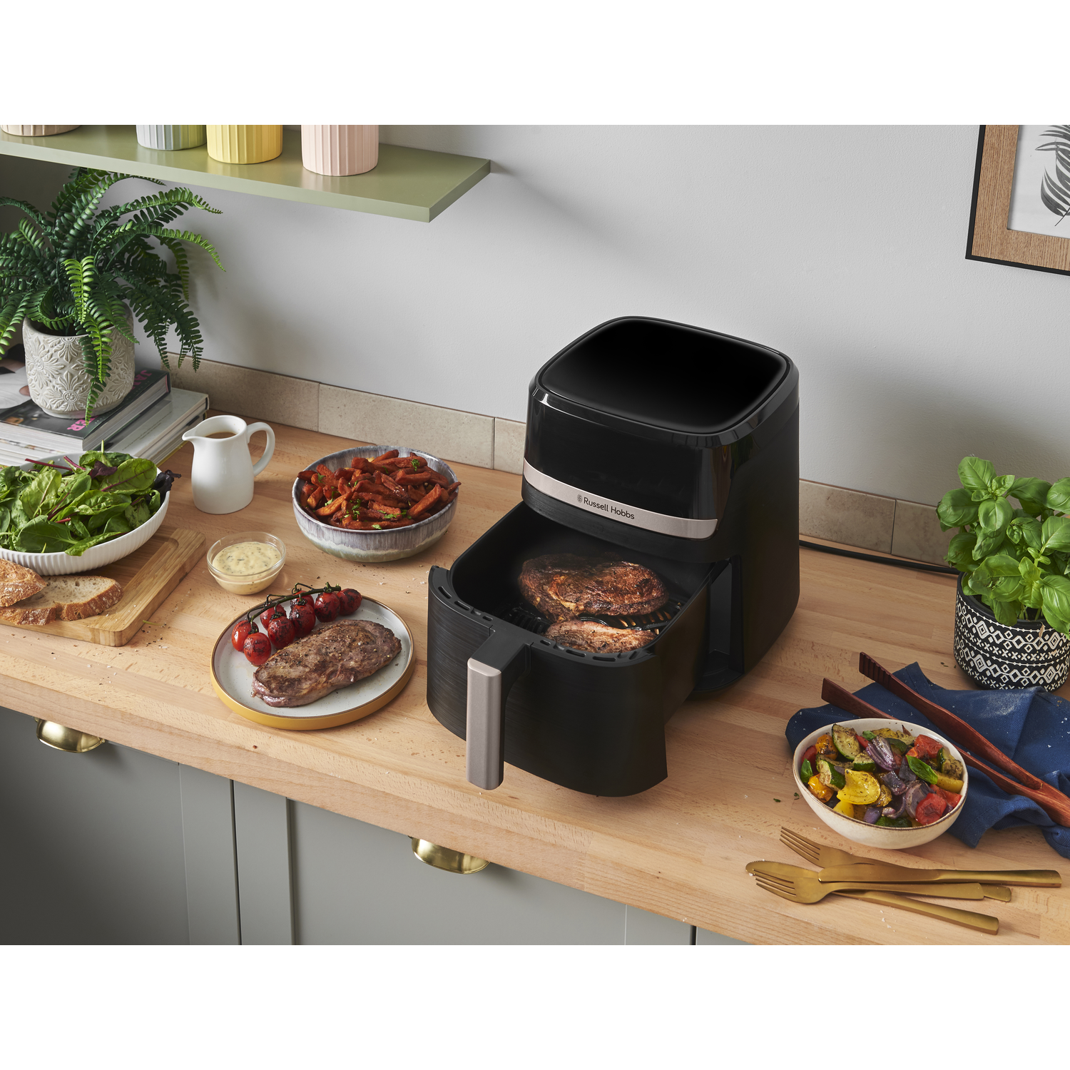 A black air fryer with food inside sits on a wooden counter, surrounded by cooked vegetables, steak, and fresh herbs.