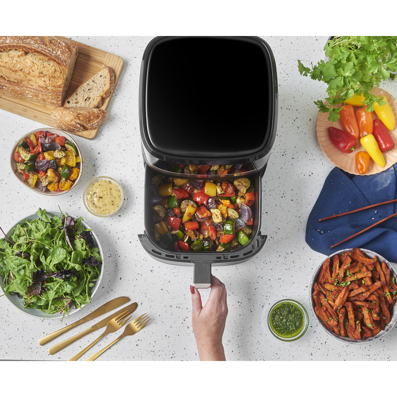 An air fryer with roasted vegetables, salad, bread, peppers, fries, and sauces on a white surface. A hand pulls out the fryer basket.