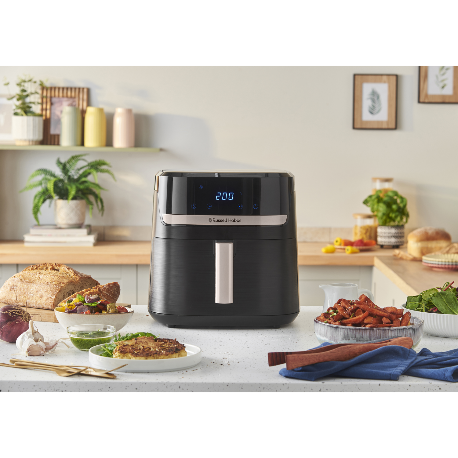 Zwarte airfryer op een aanrecht met eten. Brood, salade en andere gerechten zijn ook zichtbaar.