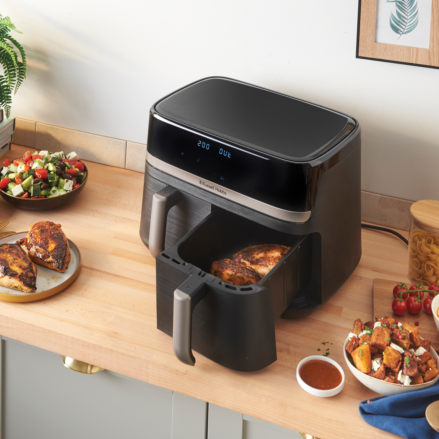 Black air fryer with food, a bowl of salad, and other food items on a kitchen counter.