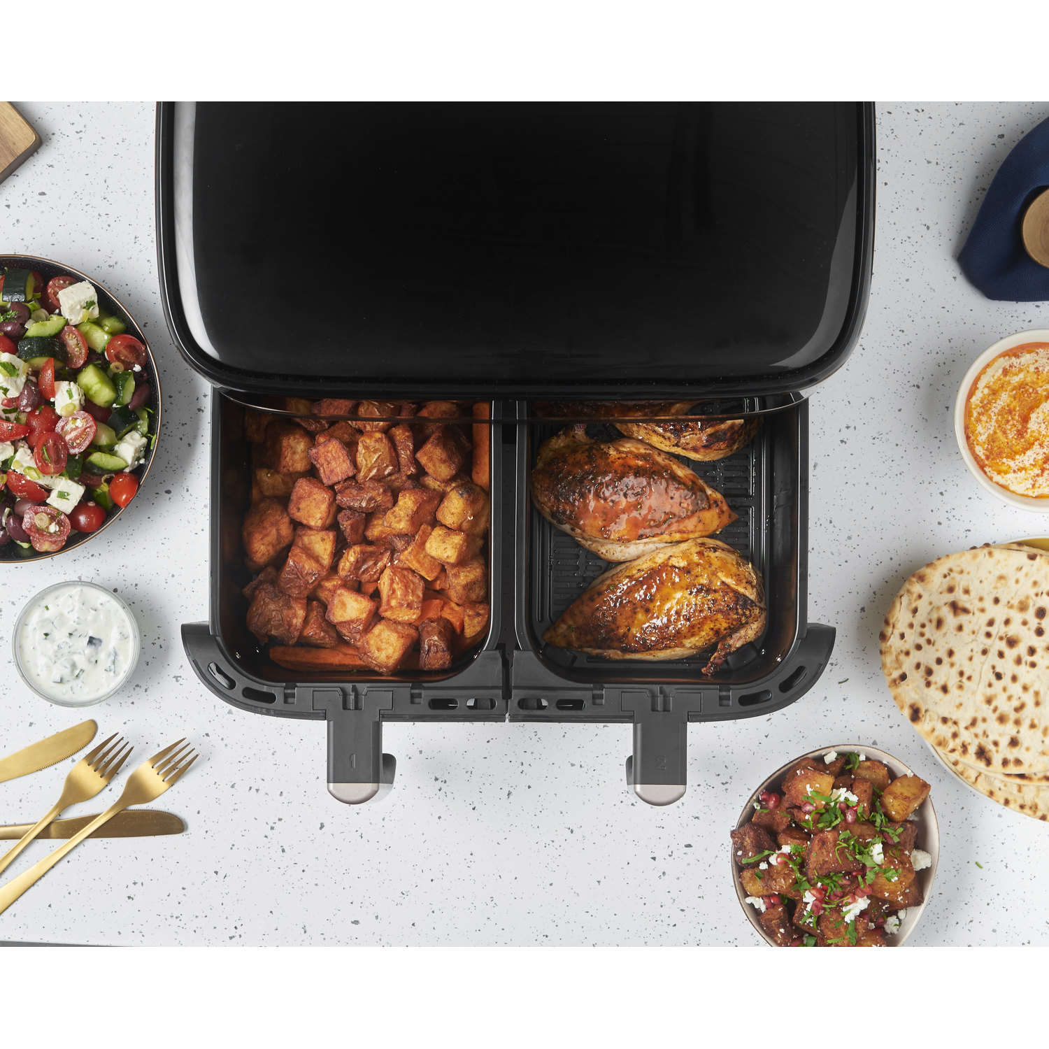 An air fryer with potatoes, chicken, and side dishes on a white surface. Gold utensils and food items are present.