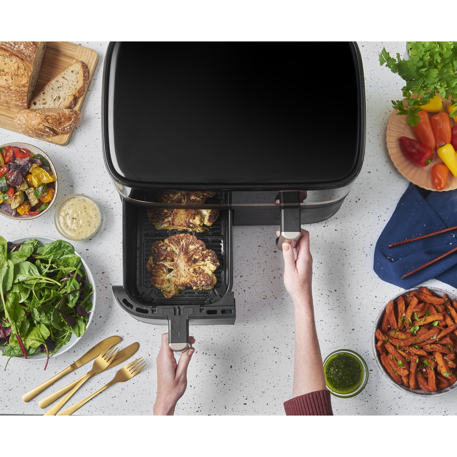 Air fryer with cooked food inside and ingredients around. Hands are holding the fryer.