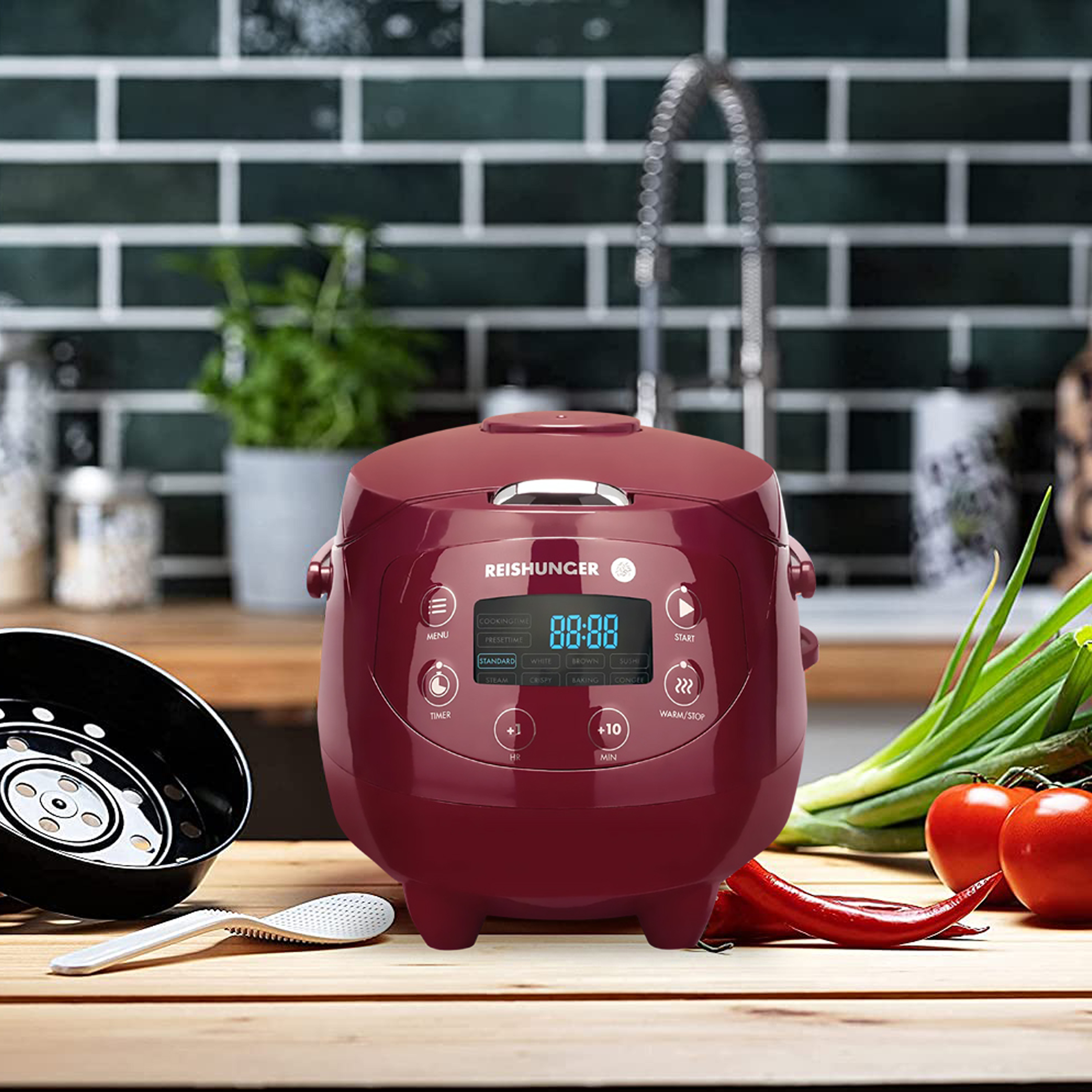 Roter Reiskocher mit Display und Knöpfen auf Holztisch, Gemüse im Hintergrund.
