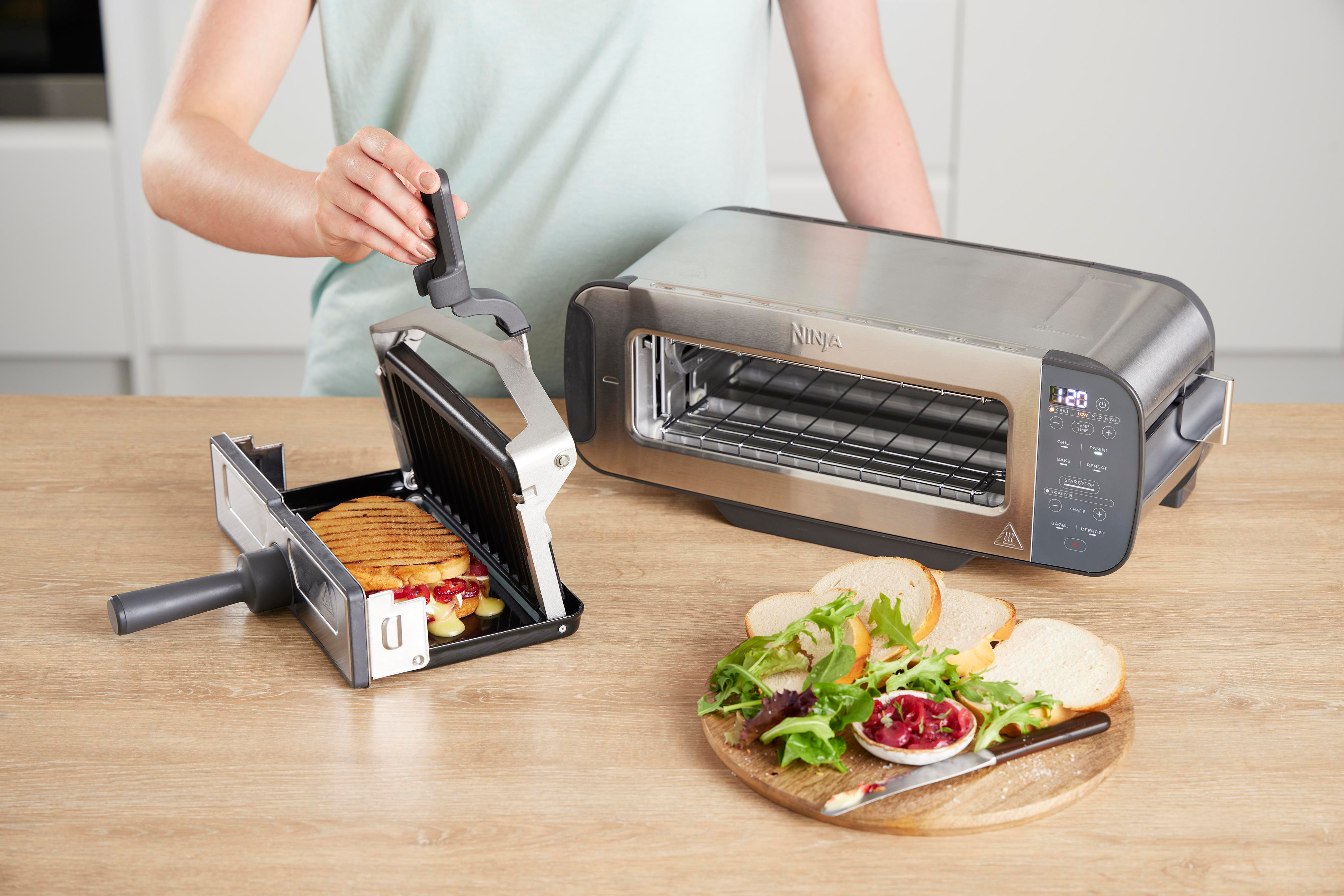 Eine Person benutzt einen Ninja-Sandwichmaker neben einem Toaster. Essen und Zutaten auf einem Tisch.