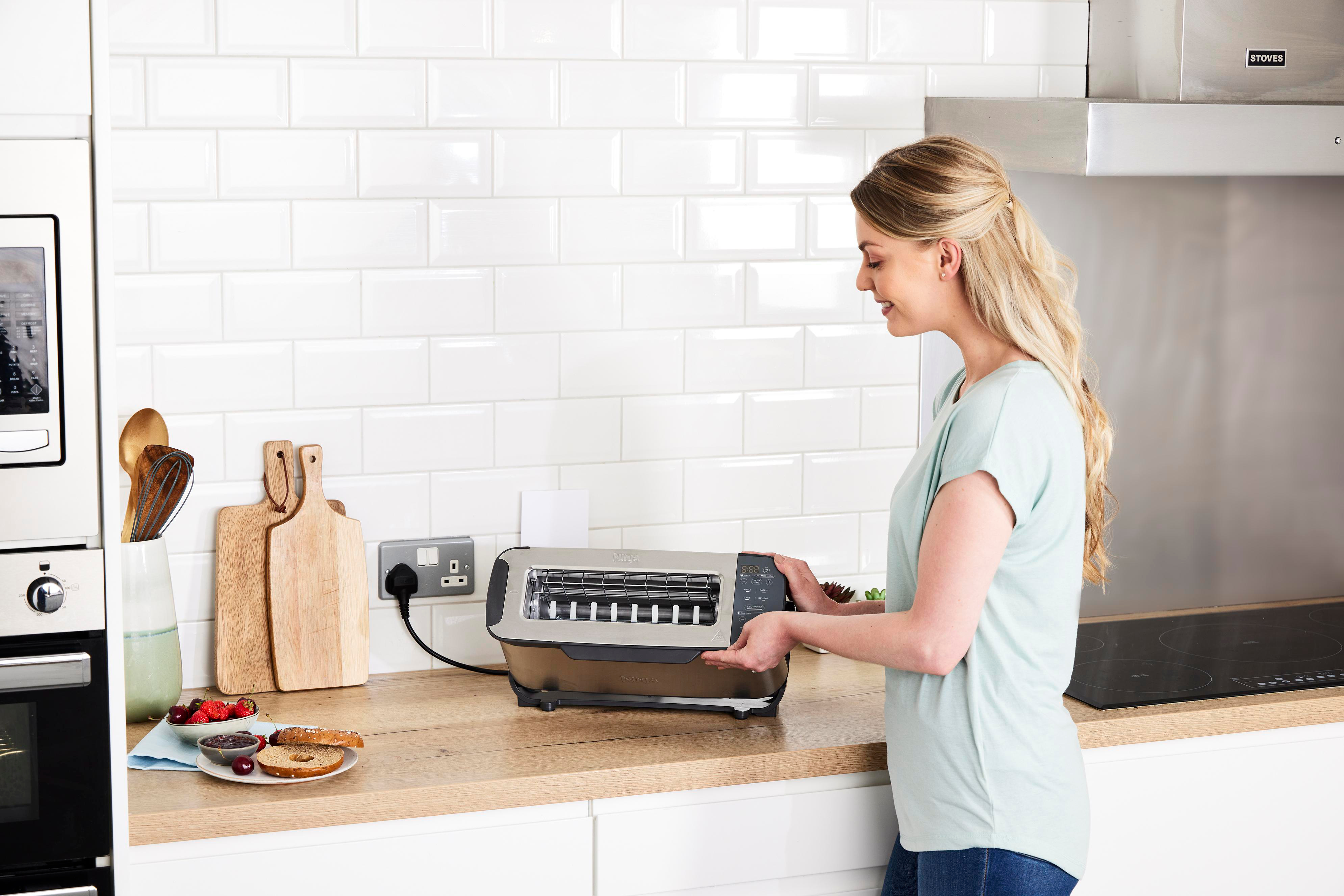 Frau in Küche schaut Toaster an, Brot und Marmelade daneben.