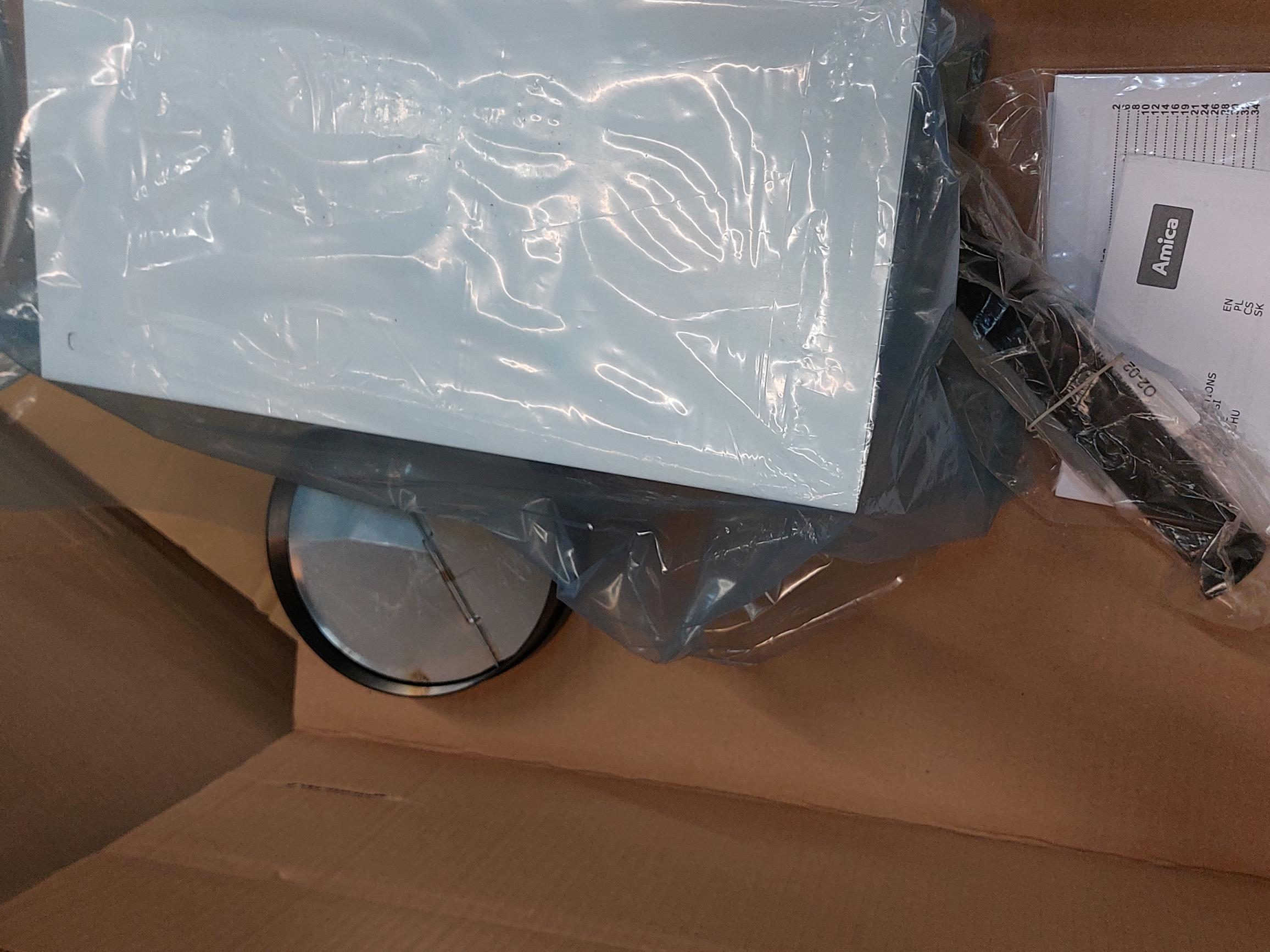 Rozpakowane białe urządzenie w kartonowym pudełku, owinięte przezroczystym plastikiem.