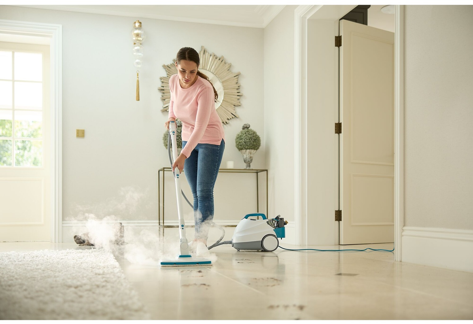 Una donna pulisce un pavimento chiaro con un pulitore a vapore. Uno specchio e una porta sono sullo sfondo.