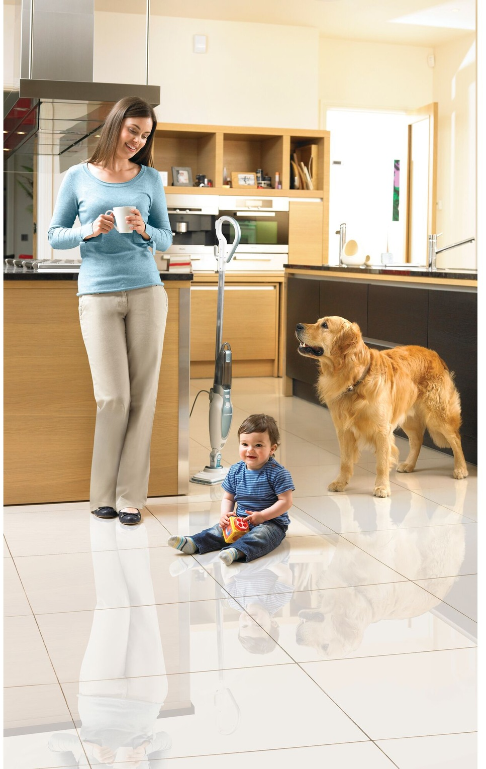 Una donna, un bambino e un cane in una cucina luminosa. La donna tiene una tazza. Il bambino è seduto per terra.