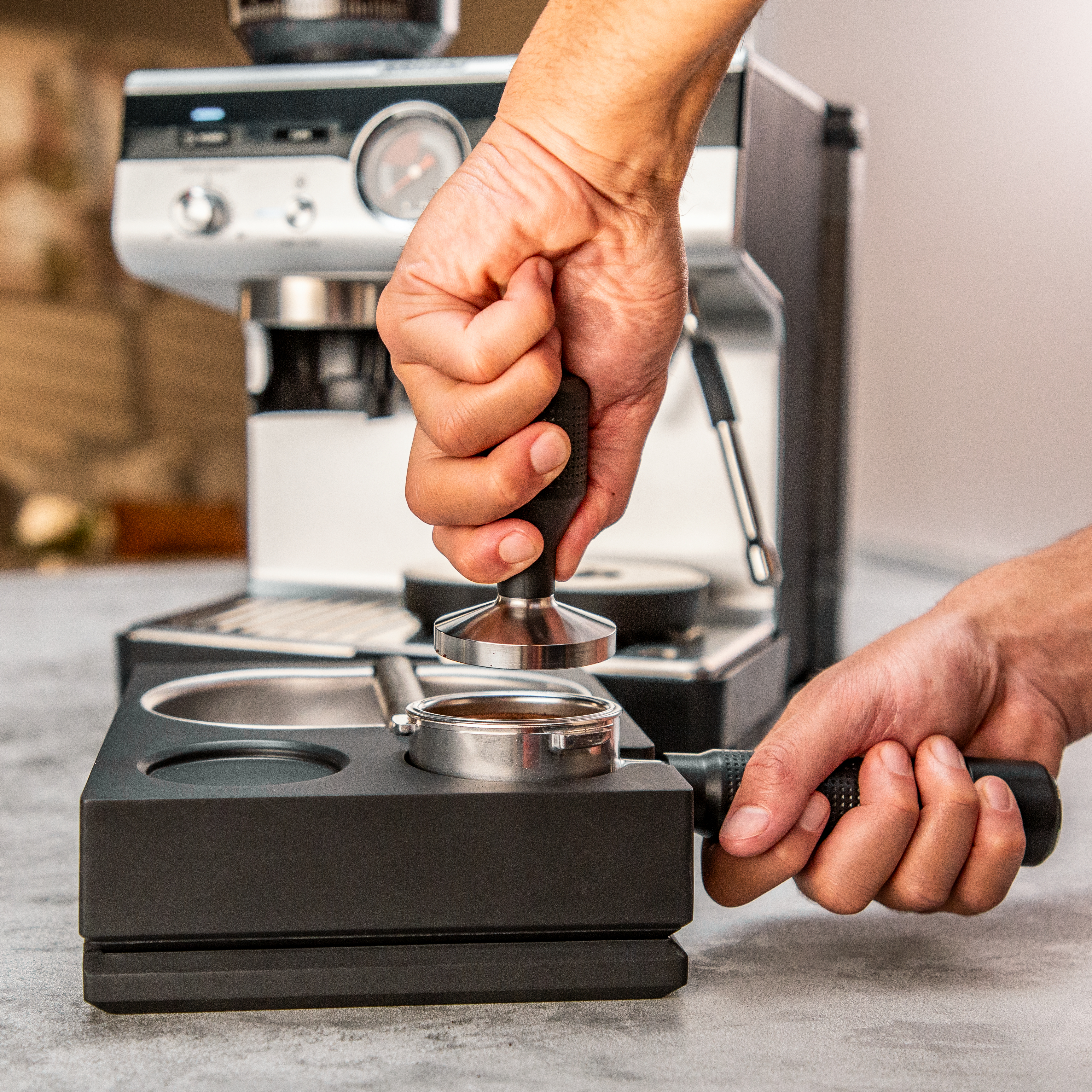 Hände, die Kaffeemehl in einem Siebträger mit einem Tamper pressen, Kaffeemaschine im Hintergrund.