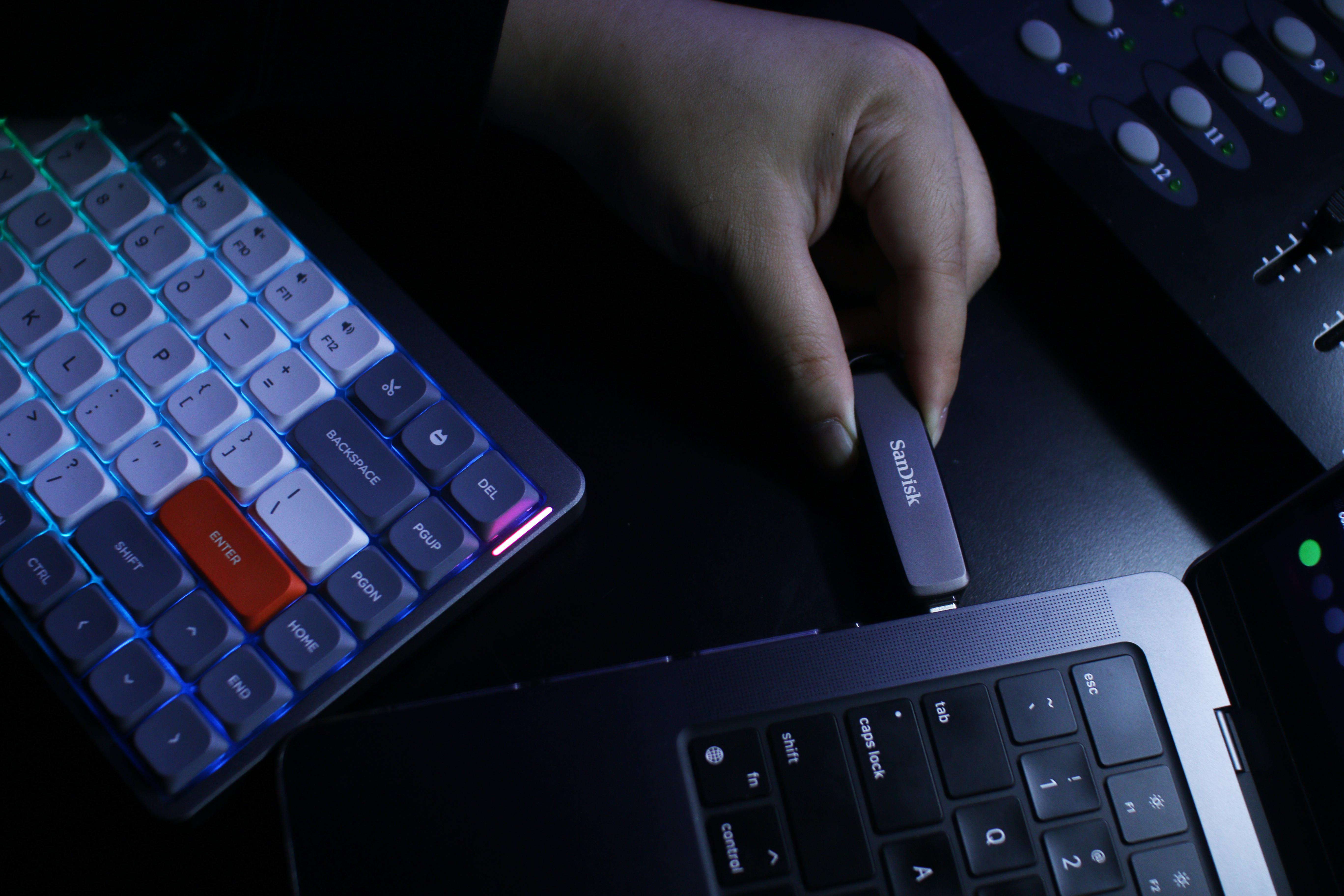 Eine Hand, die einen silbernen USB-Stick in einen Laptop neben einer Tastatur und anderen Geräten steckt.
