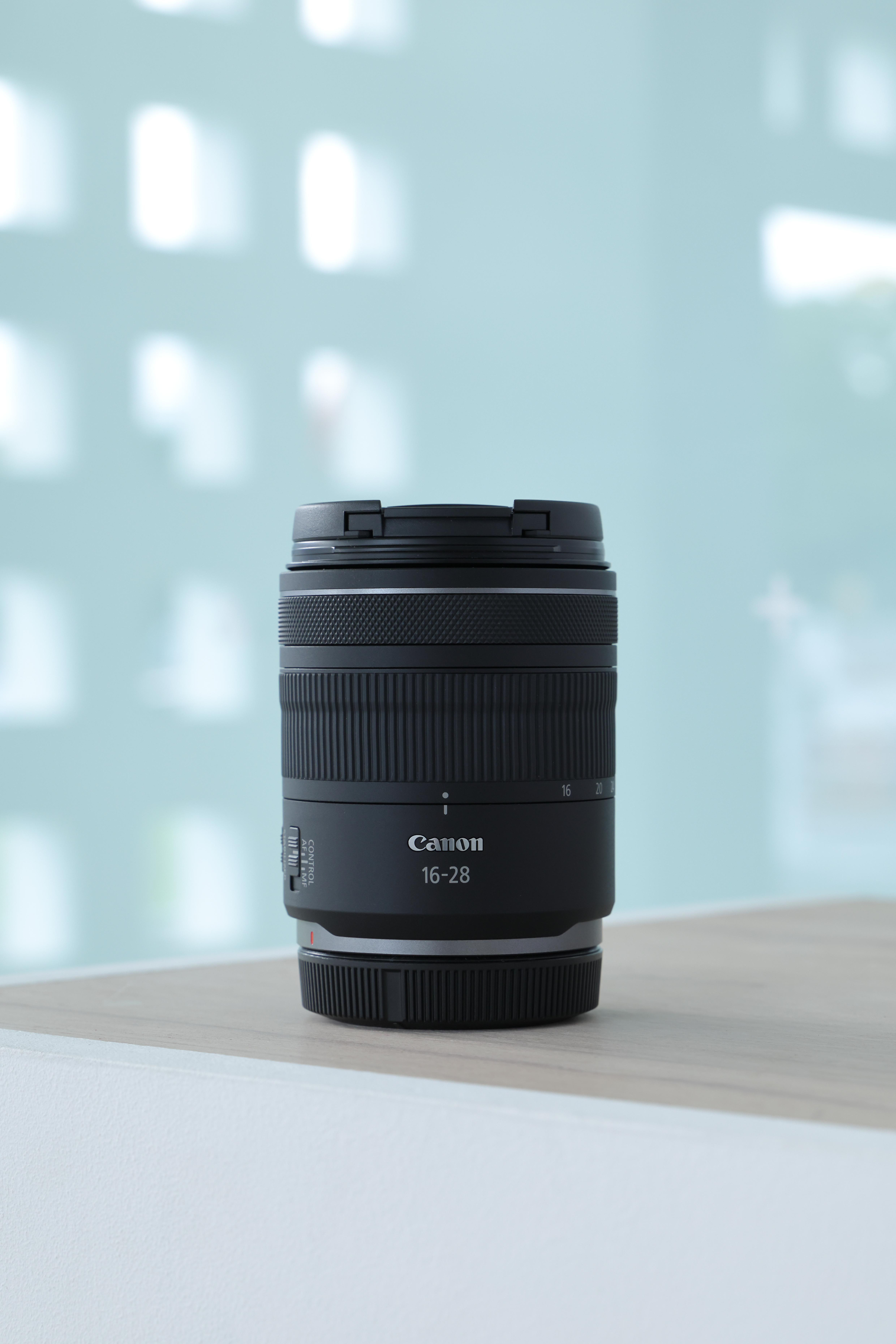 Canon 16-28mm Objektiv auf einer Holzoberfläche, mit hellblauem Hintergrund.