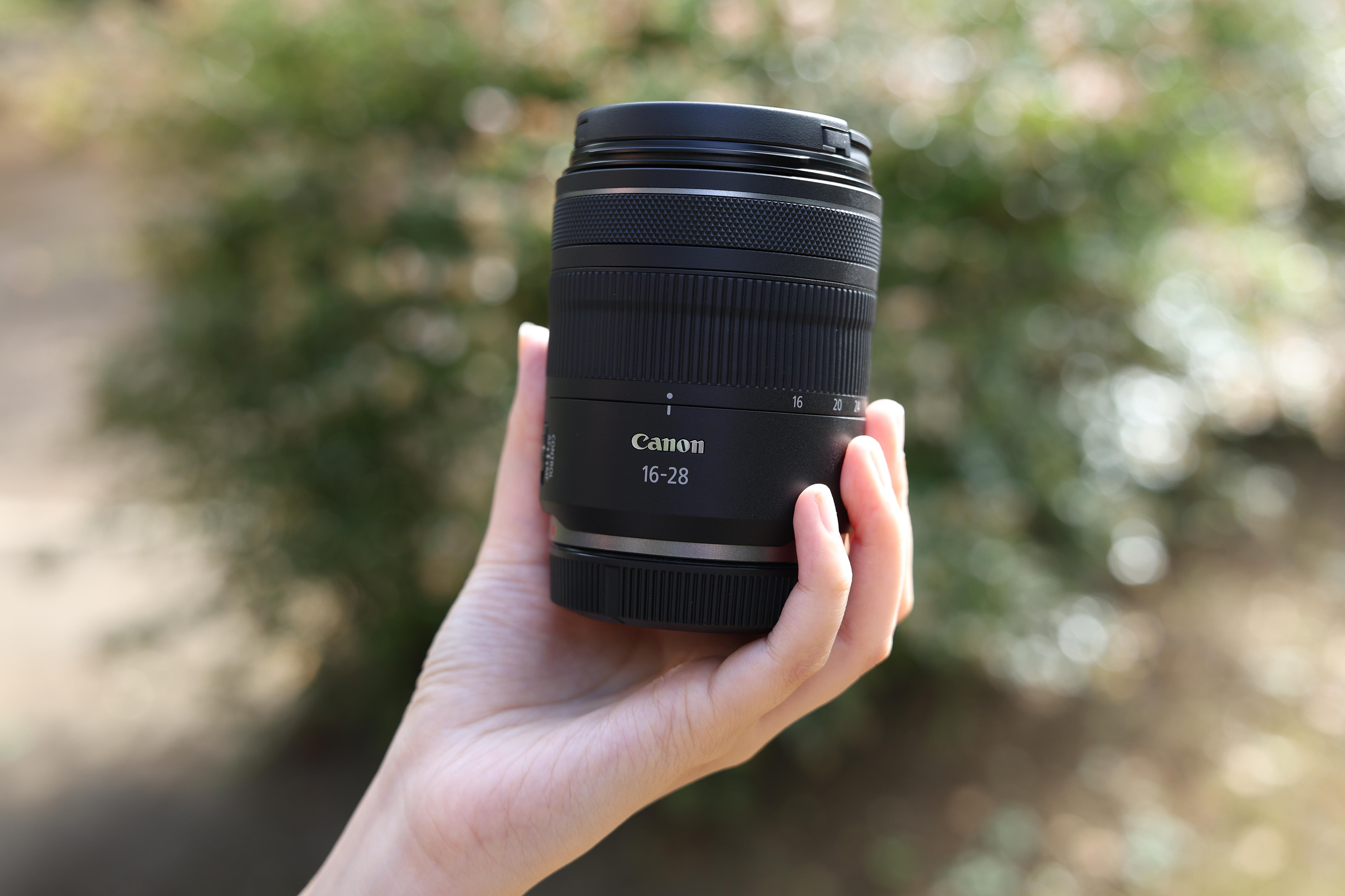 Hand, die ein schwarzes Kameraobjektiv hält, auf dem Canon 16-28 steht.