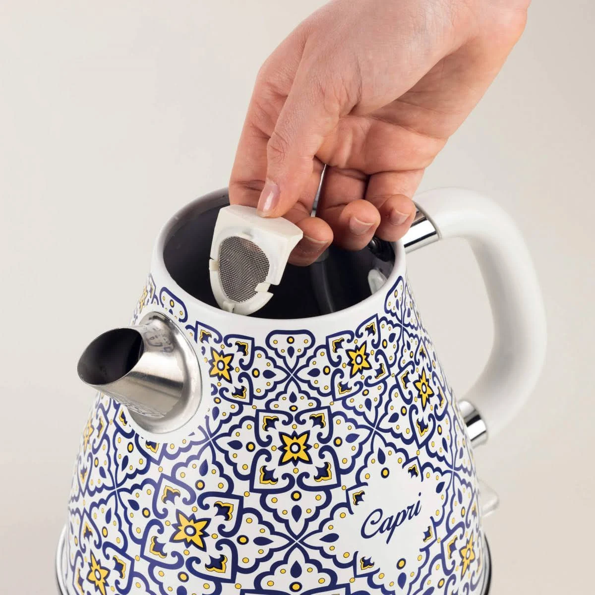 Mano colocando un filtro dentro de una tetera decorada. La tetera tiene un patrón azul, amarillo y blanco. La palabra Capri está en ella.