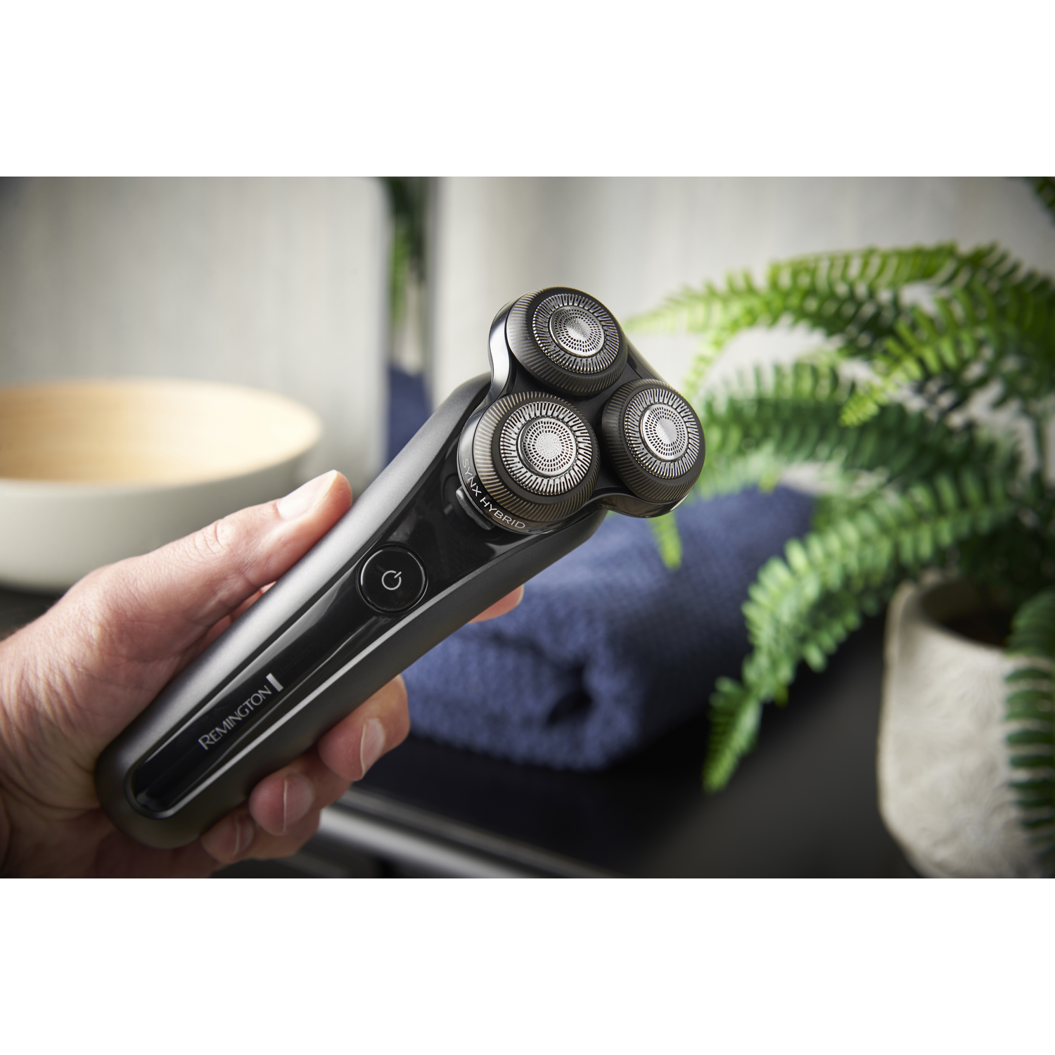 A man holding a black and silver Remington rotary shaver near a blue towel and green plant.