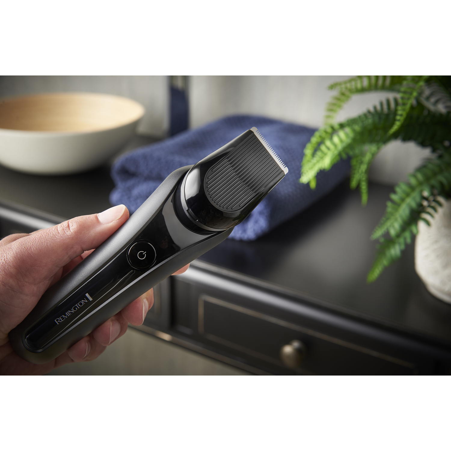 Hand holding a Remington beard trimmer. Blue towel and plant in background.