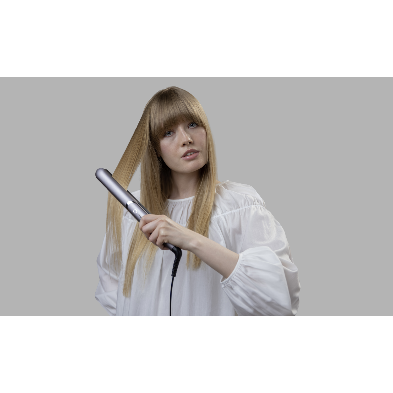 Woman with blonde hair using a hair straightener. Gray background.
