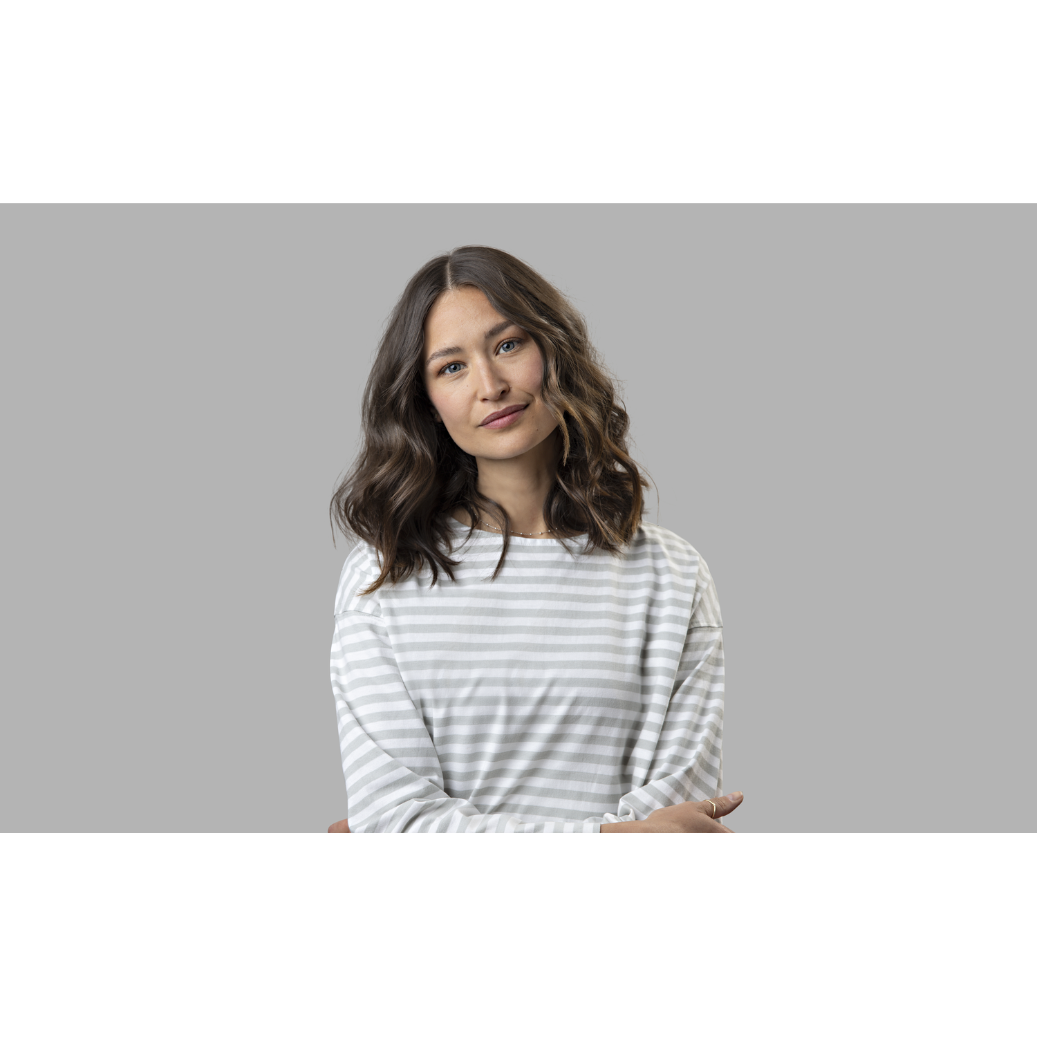 A woman with wavy brown hair, wearing a striped shirt, poses with a neutral expression on a gray background.