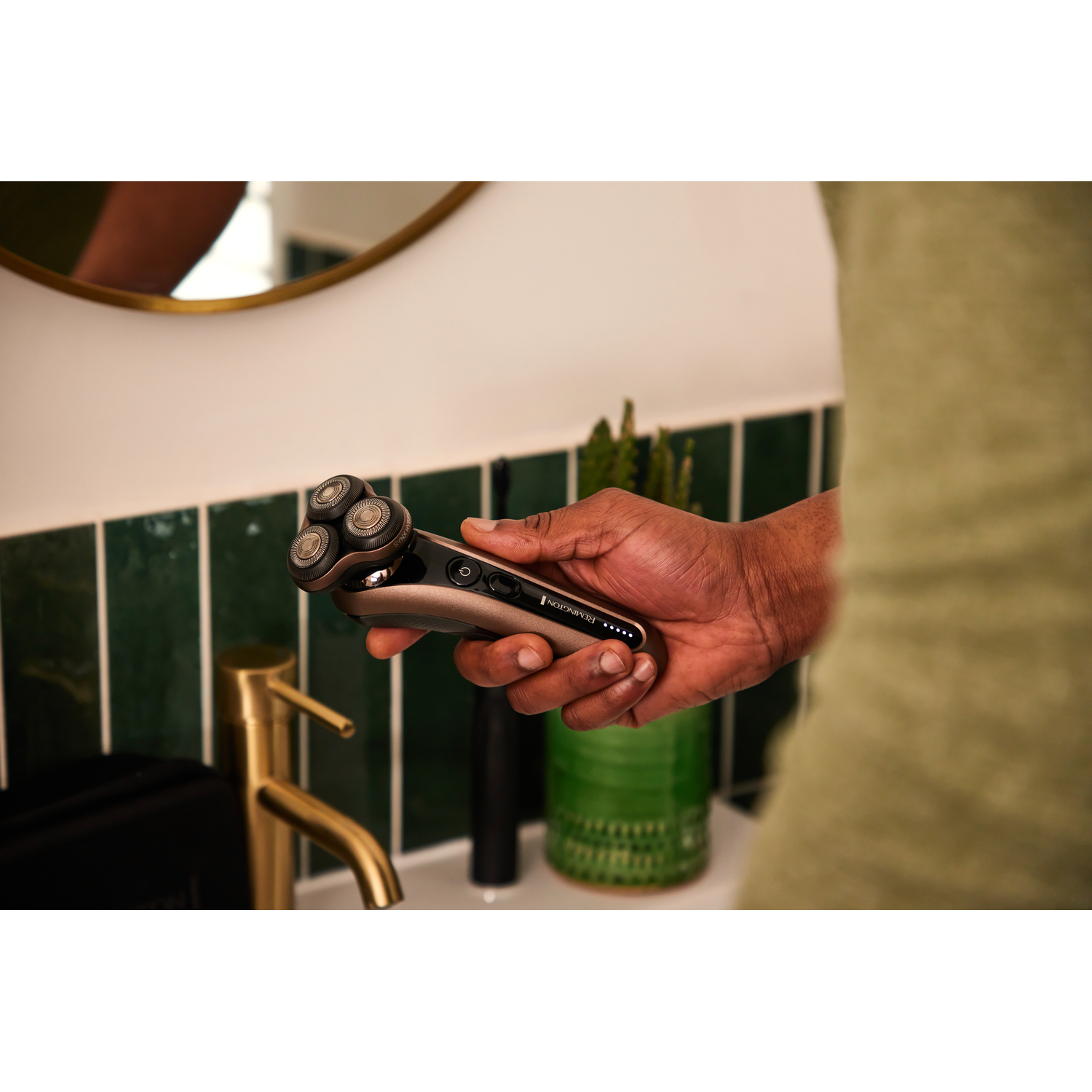 Man holding a silver and brown shaver in front of a mirror, with a sink and a green wall.