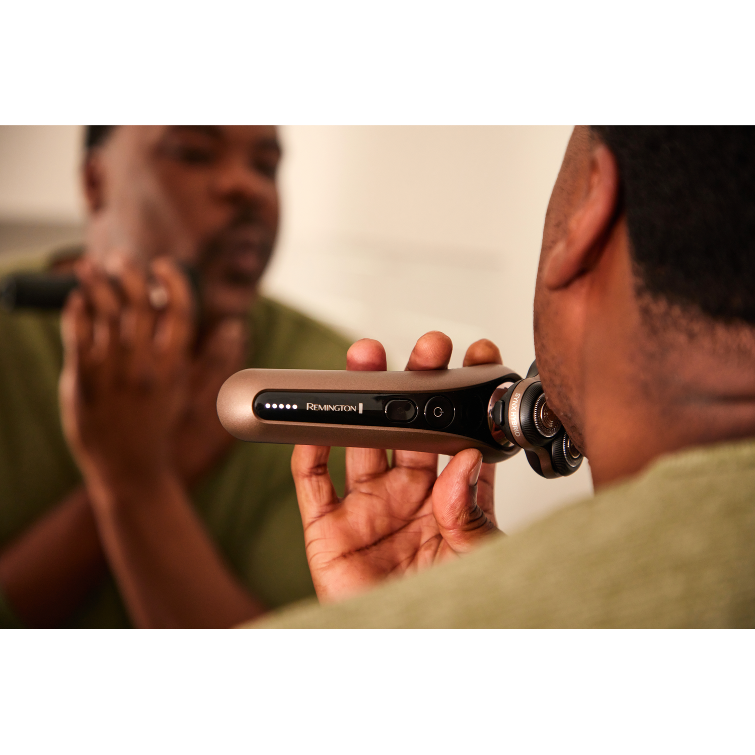 A man shaves his face in a mirror with a Remington shaver. The shaver is gold-colored.