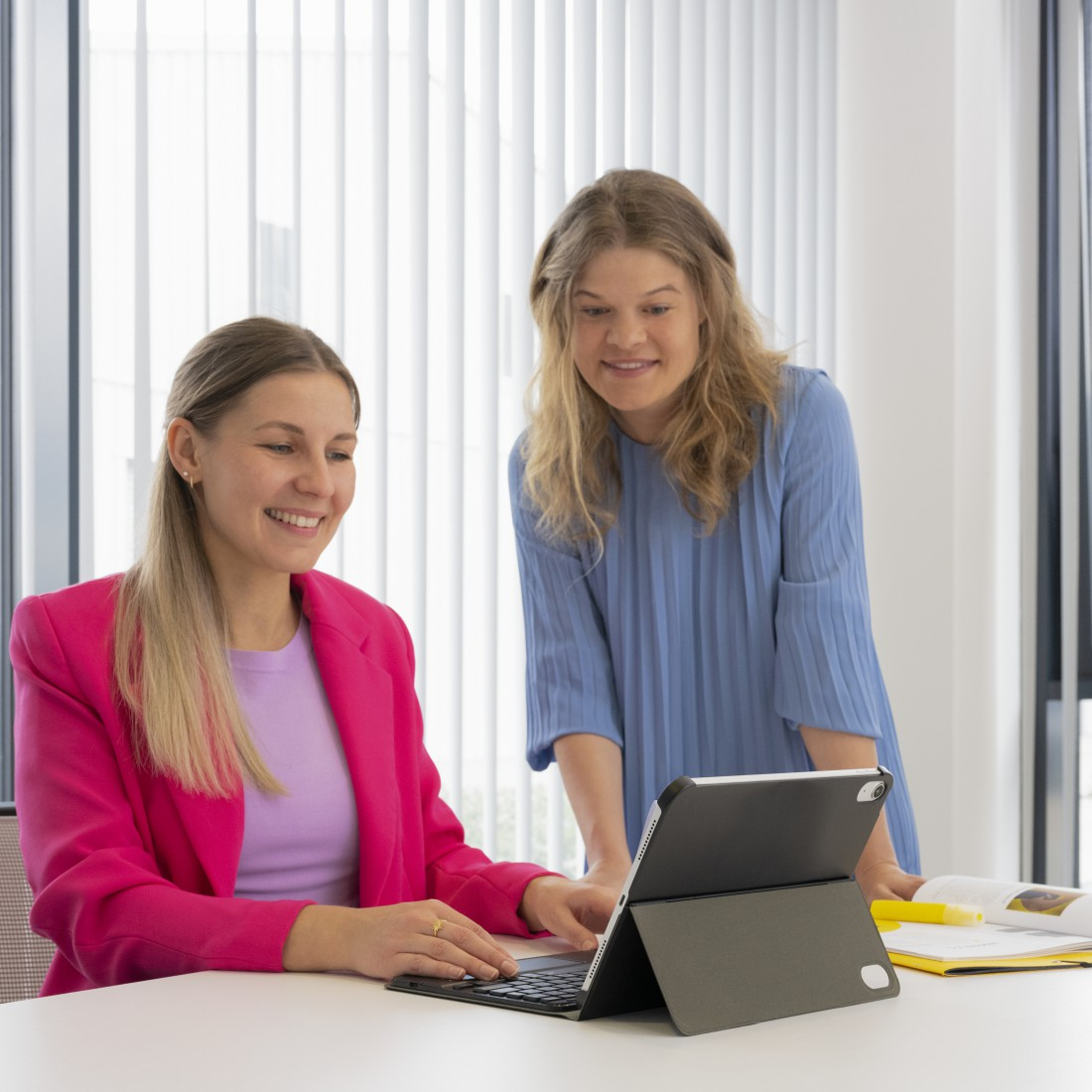 Zwei Frauen arbeiten zusammen und betrachten ein Tablet mit angeschlossener Tastatur. Ein Tisch ist vorhanden.