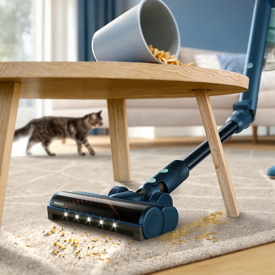 Niebieski odkurzacz na dywanie z rozsypanymi okruchami. Kot przechodzi obok stołu.