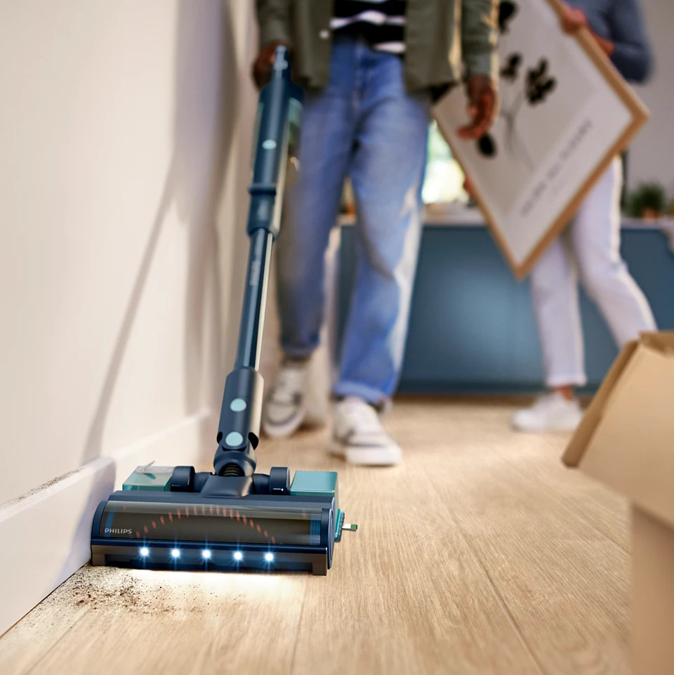Osoba odkurzająca podłogę w pobliżu ściany, inne osoby i przedmioty w tle.