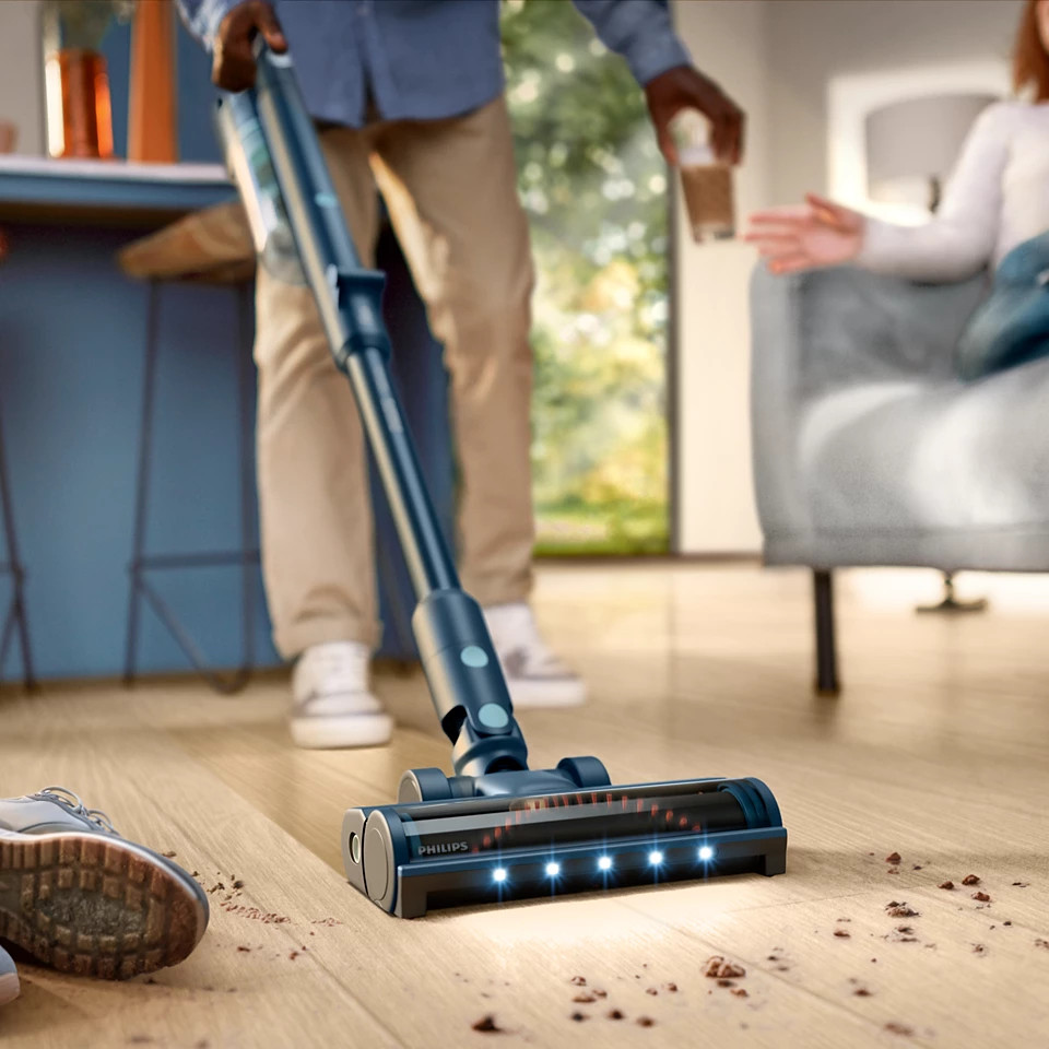 Osoba używająca niebiesko-czarnego odkurzacza na drewnianej podłodze. Widać drugą osobę i filiżankę kawy.