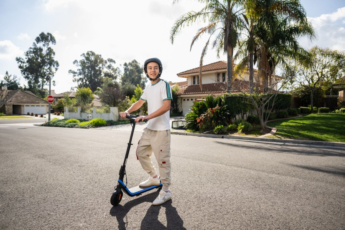 Junge auf einem Roller, mit Helm, auf einer Straße mit Bäumen und Häusern im Hintergrund.