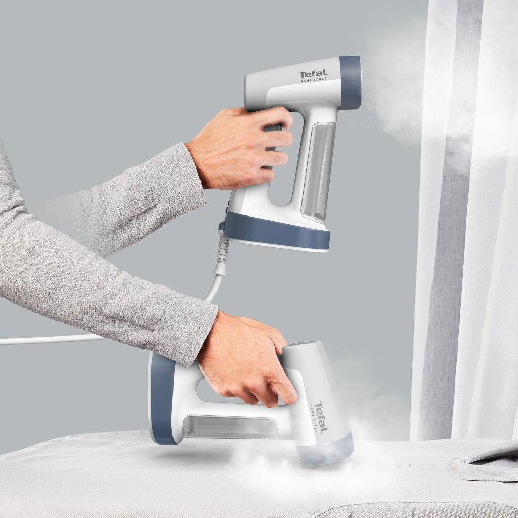 Dłonie trzymają żelazko parowe Tefal, prasują poduszkę, szare tło.