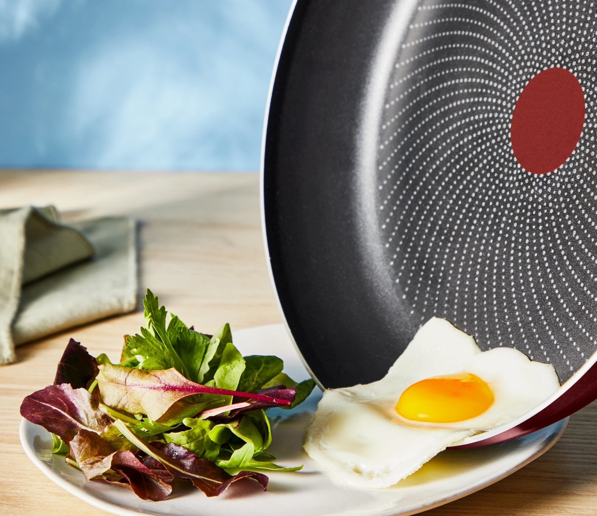Jajko na patelni, serwowane z sałatką. Biały talerz, drewniany stół, niebieskie tło.