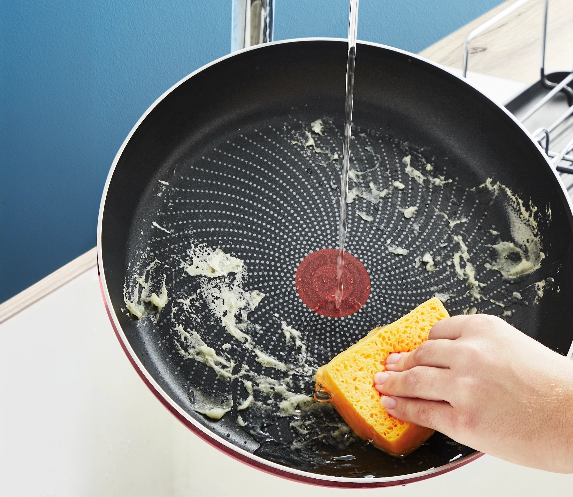 Ręka myje czerwoną patelnię. Woda spływa po niej, a żółta gąbka jest używana.