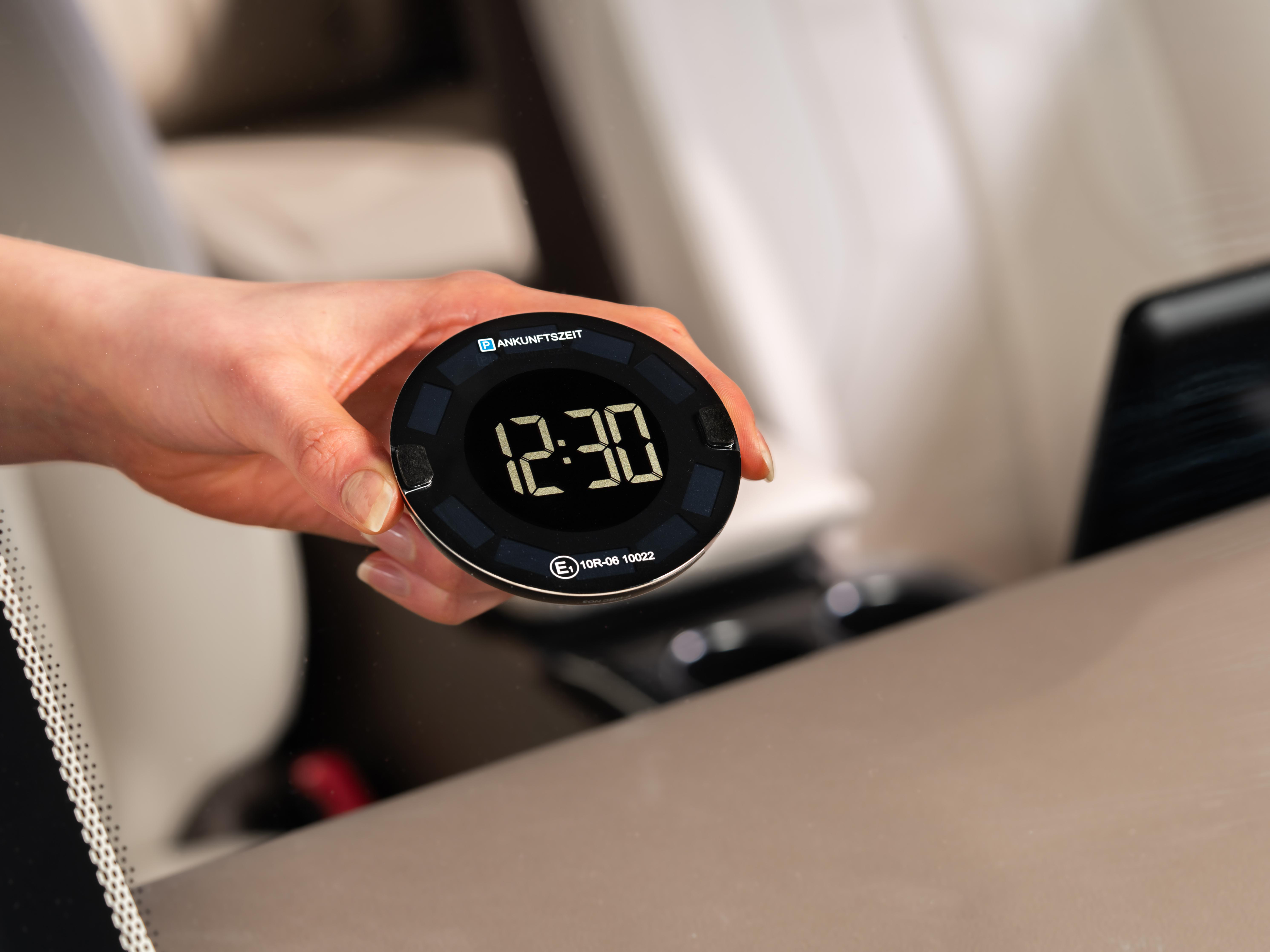 Eine Frau hält eine Autouhr, die 12:30 anzeigt, mit einem Auto-Innenraumhintergrund.