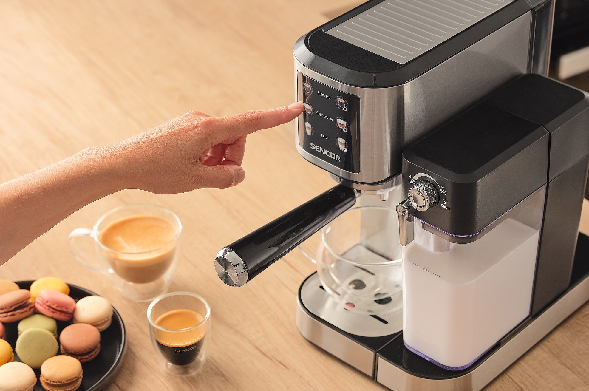 Személy keze megnyomja a cappuccino gombot egy eszpresszó gépen. Kávé és macaronok a közelben.