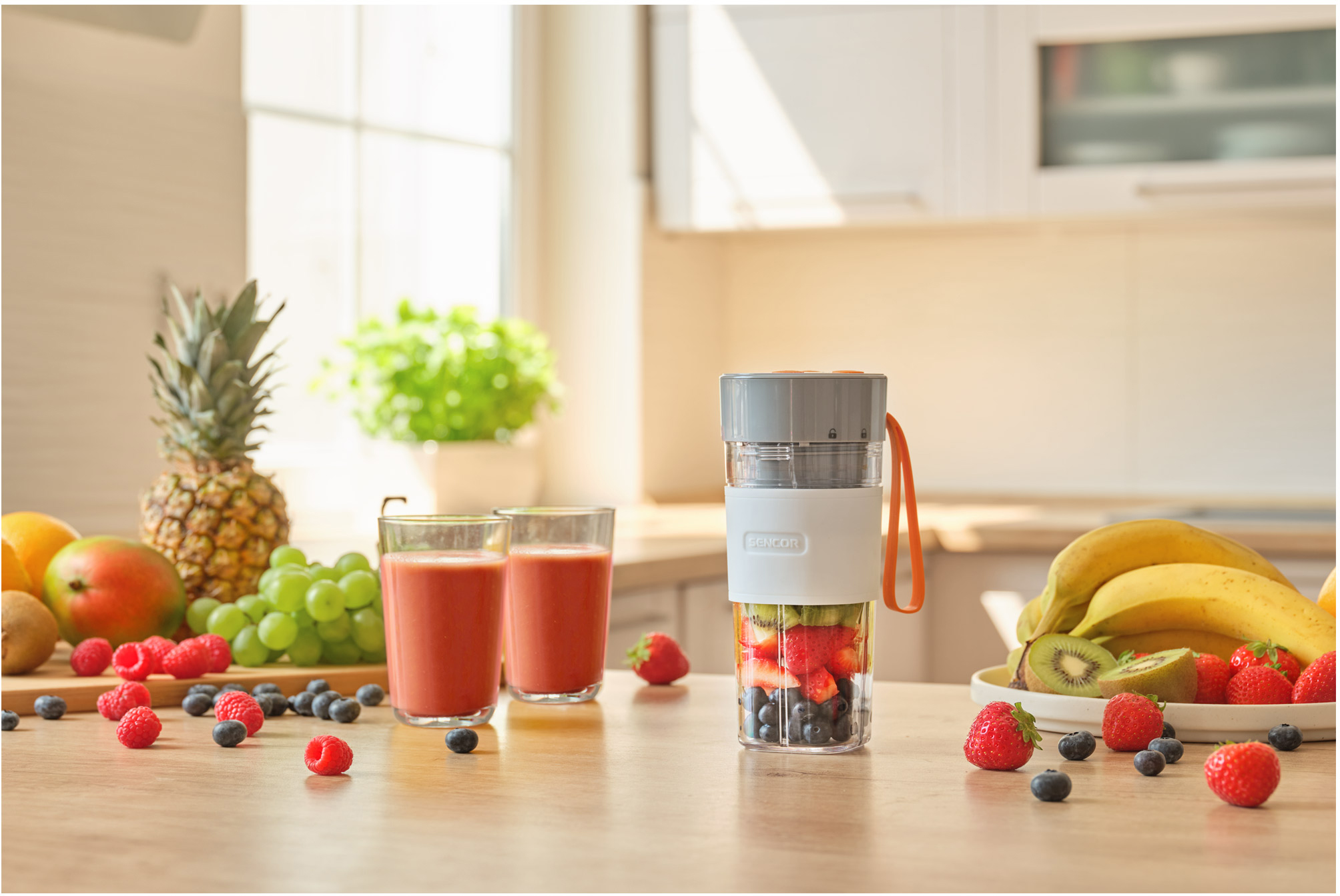 Egy turmixgép gyümölcsökkel, és két pohár smoothie egy fa asztalon. A háttér egy világos konyha.