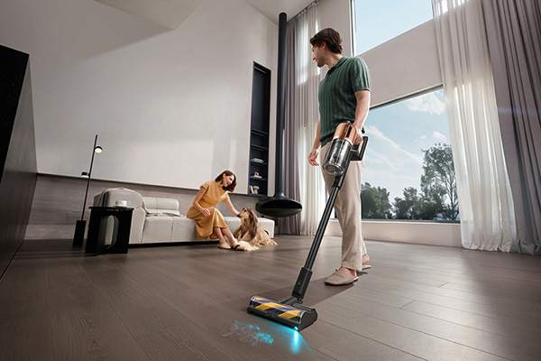 Mężczyzna odkurza drewnianą podłogę, a kobieta siedzi na kanapie z psem.