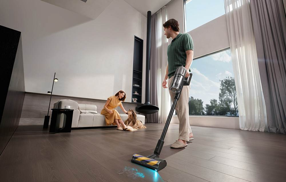 Mężczyzna odkurza podłogę nowoczesnym odkurzaczem. Kobieta na sofie z psem.
