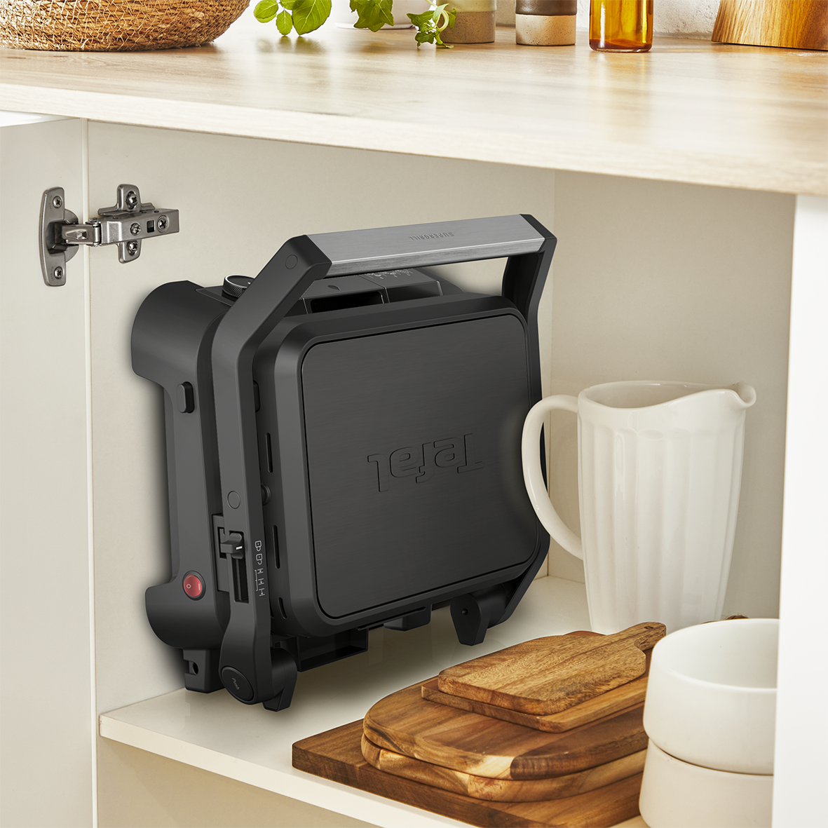 Egy fekete Tefal grill egy fehér szekrényben. Egy fehér kancsó és vágódeszkák vannak a közelben.