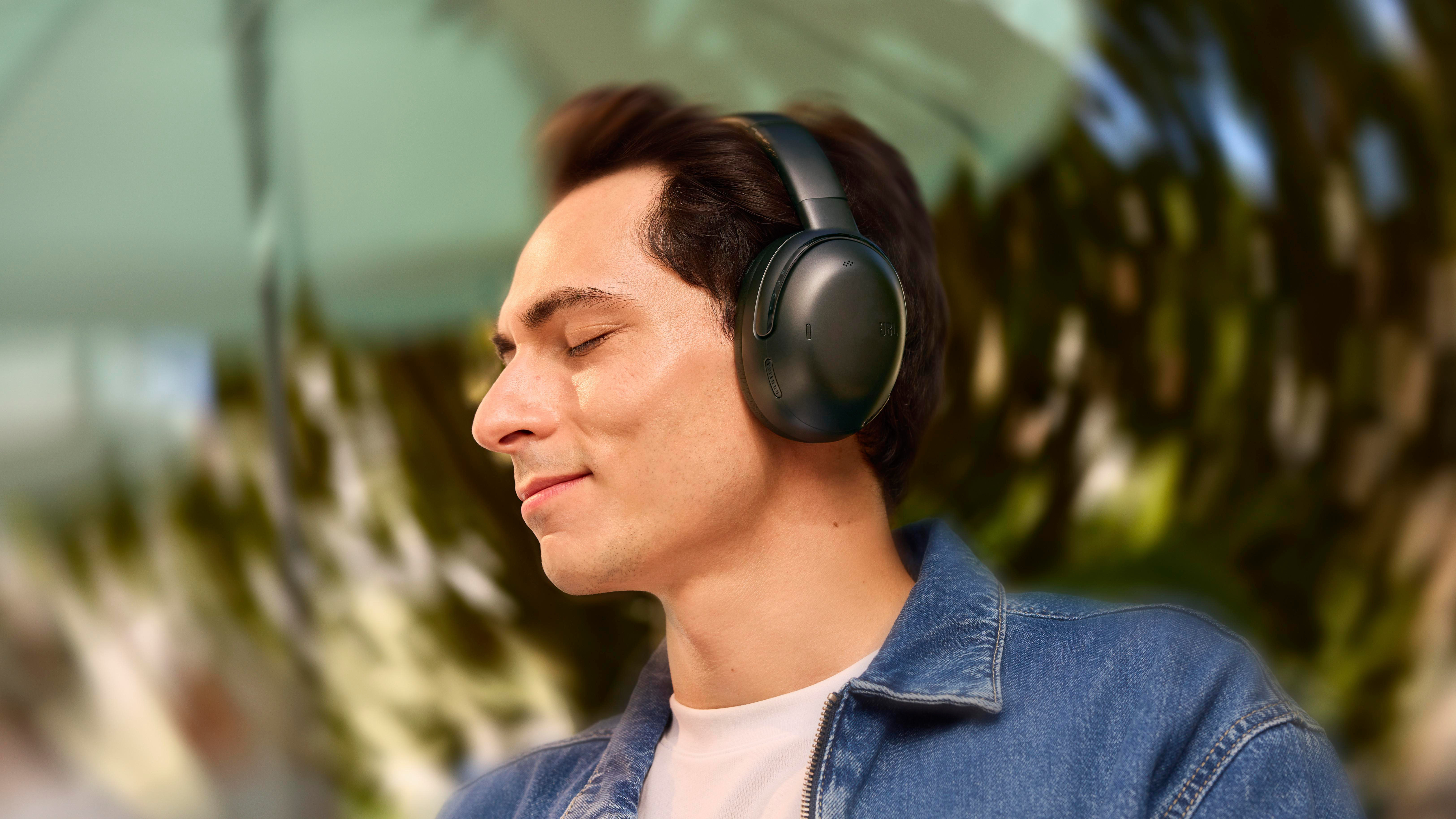 Mann mit schwarzen Kopfhörern, geschlossenen Augen, Jeansjacke, unscharfer Hintergrund.