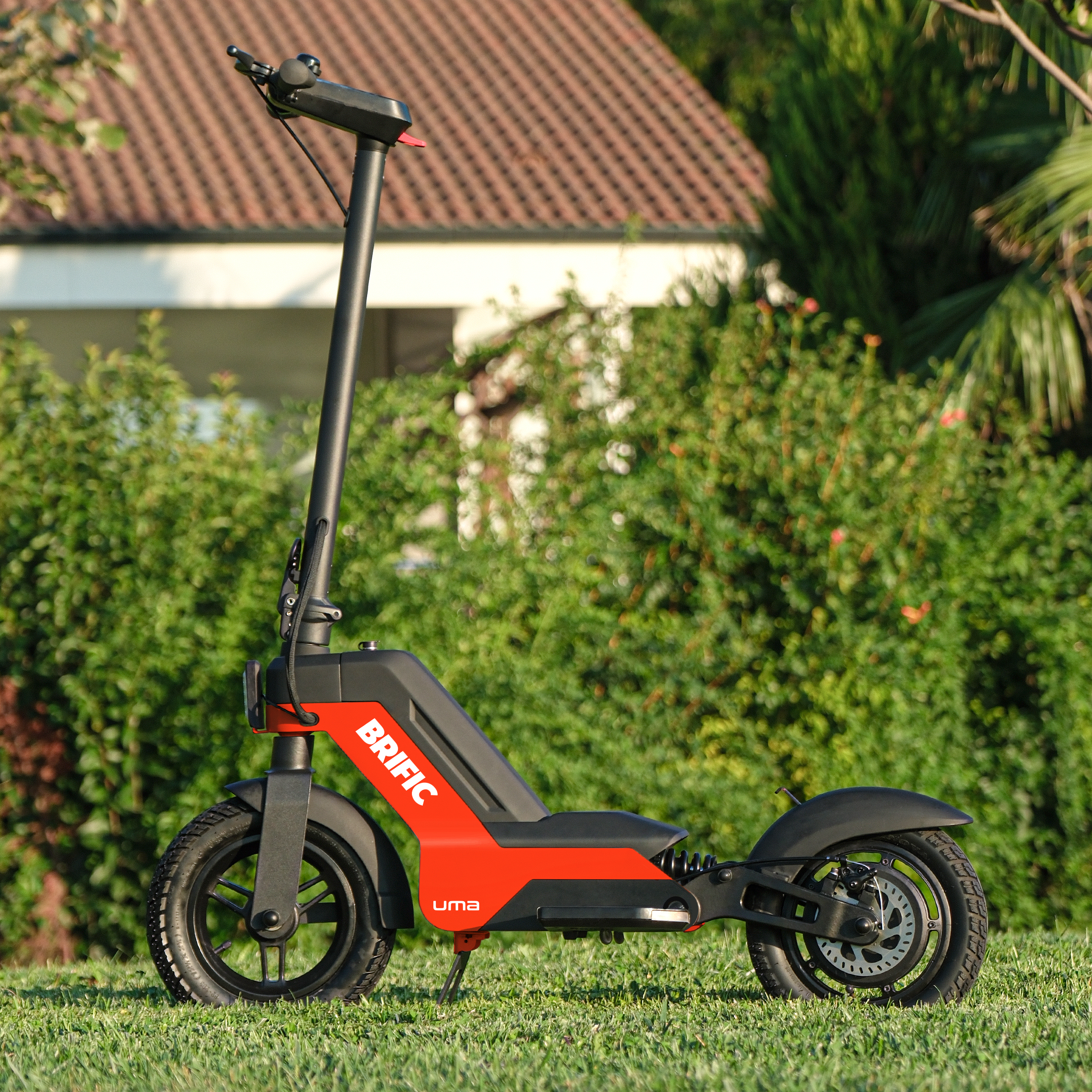 Kırmızı ve siyah bir elektrikli scooter, yeşil çimenlerin üzerinde, arka planda bir ev ile park halinde.