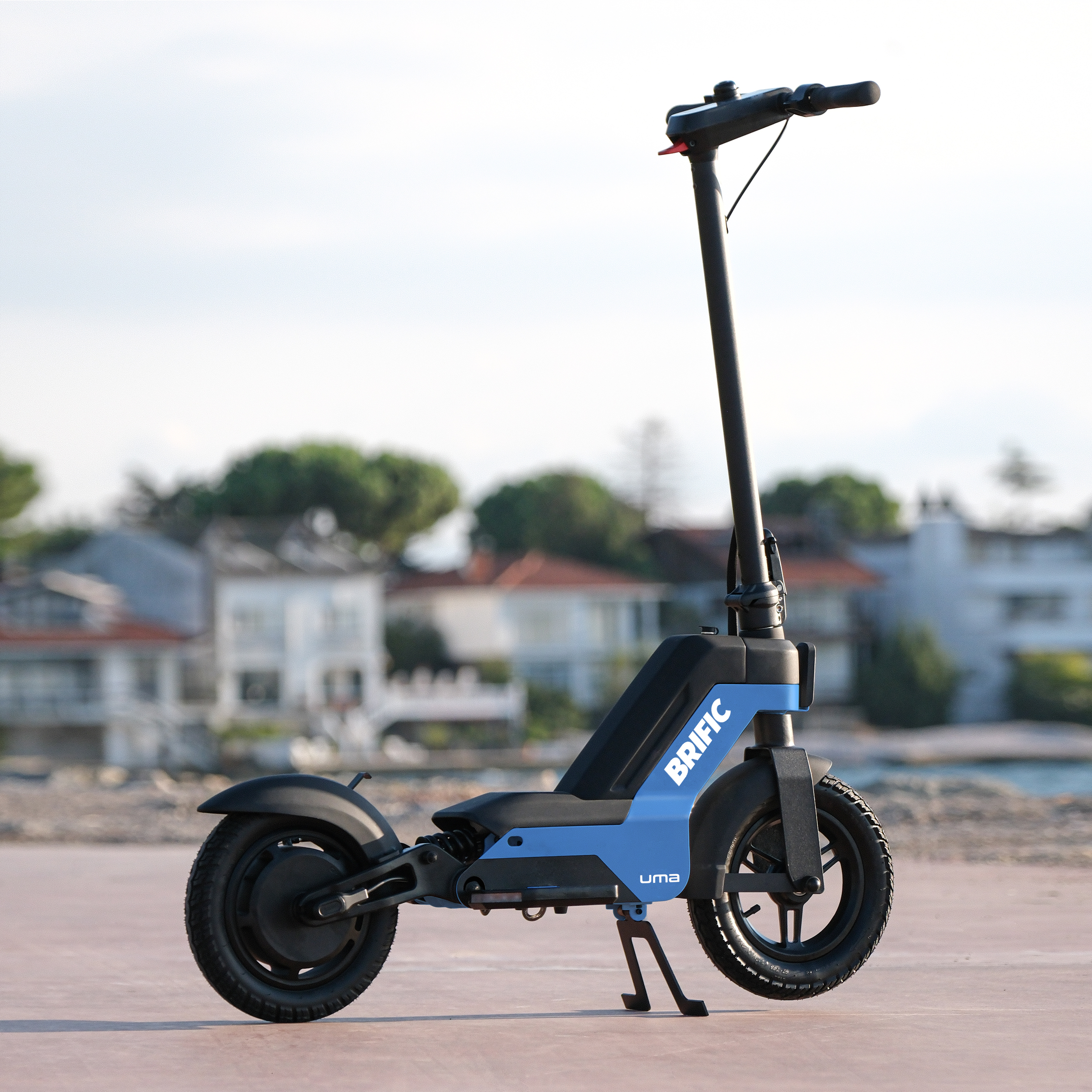 Mavi ve siyah elektrikli scooter, marka BRIFIC. Scooter, beton bir yüzeyde, arka planda doğa var.
