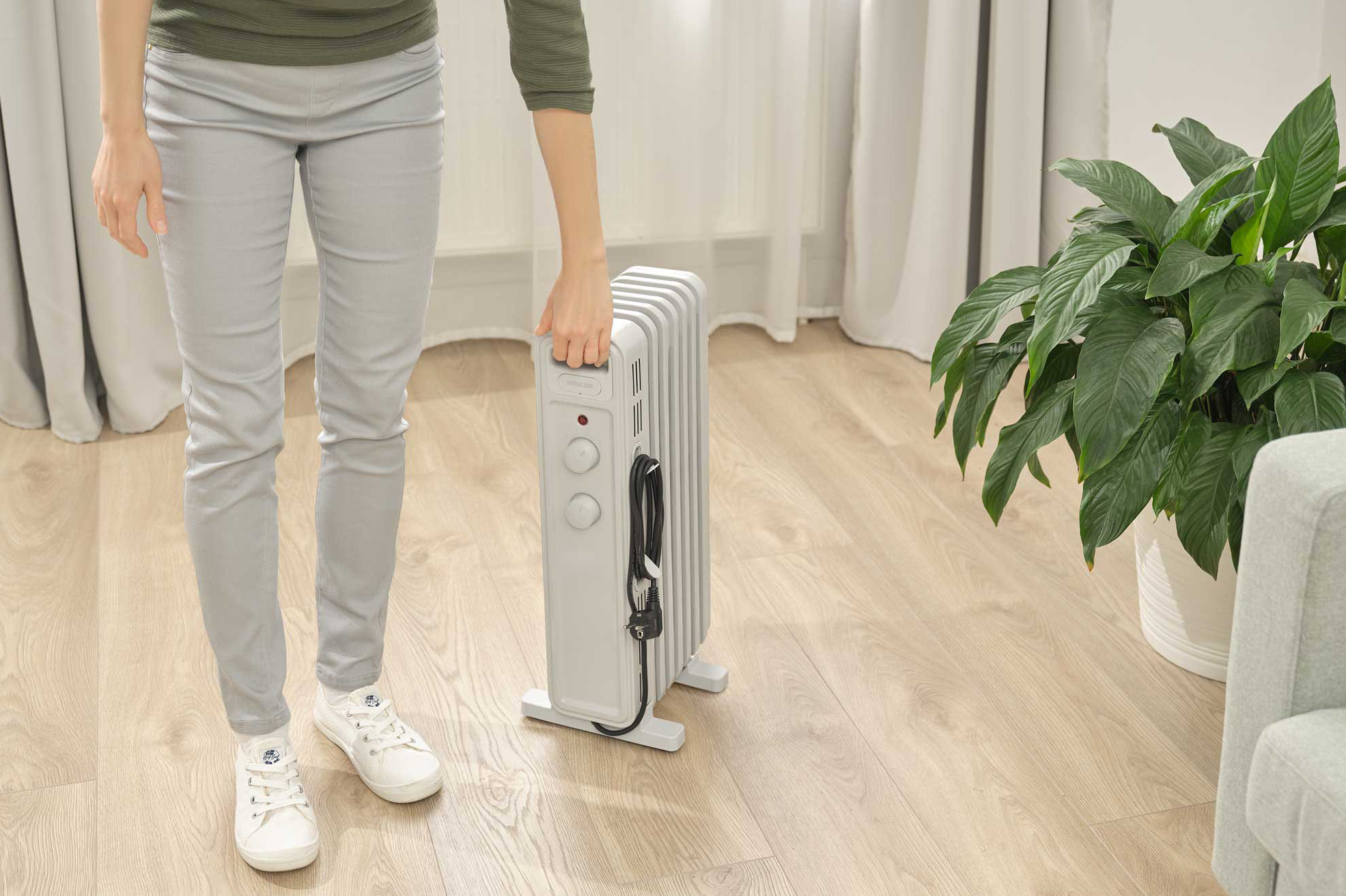 Egy személy egy szürke elektromos fűtőtestet tart egy fapadlón.