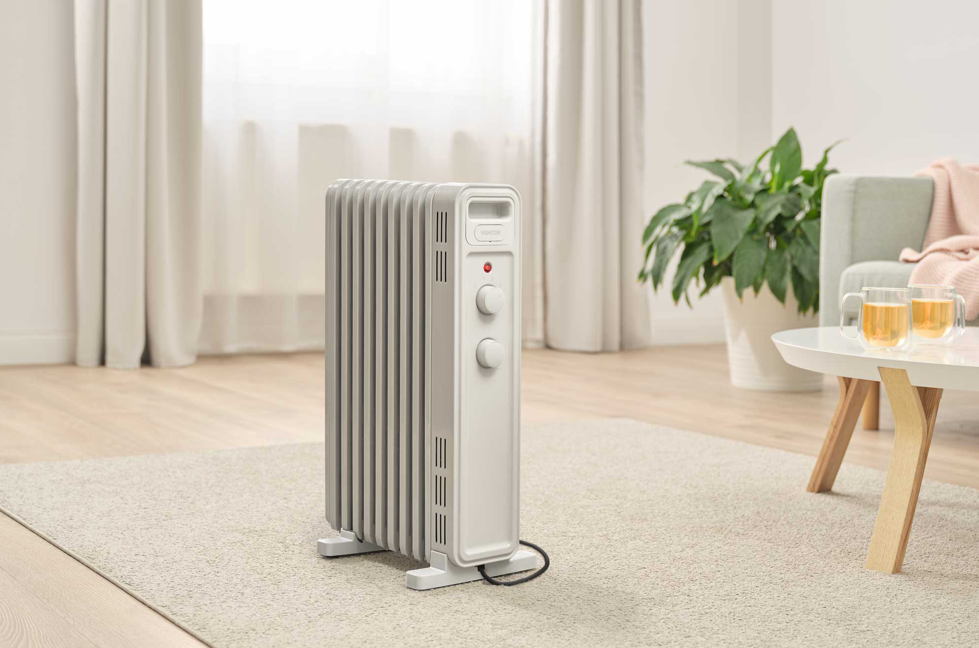 Egy fehér elektromos fűtőtest két vezérlőgombbal, egy szőnyegen egy kanapéval és egy növénnyel.