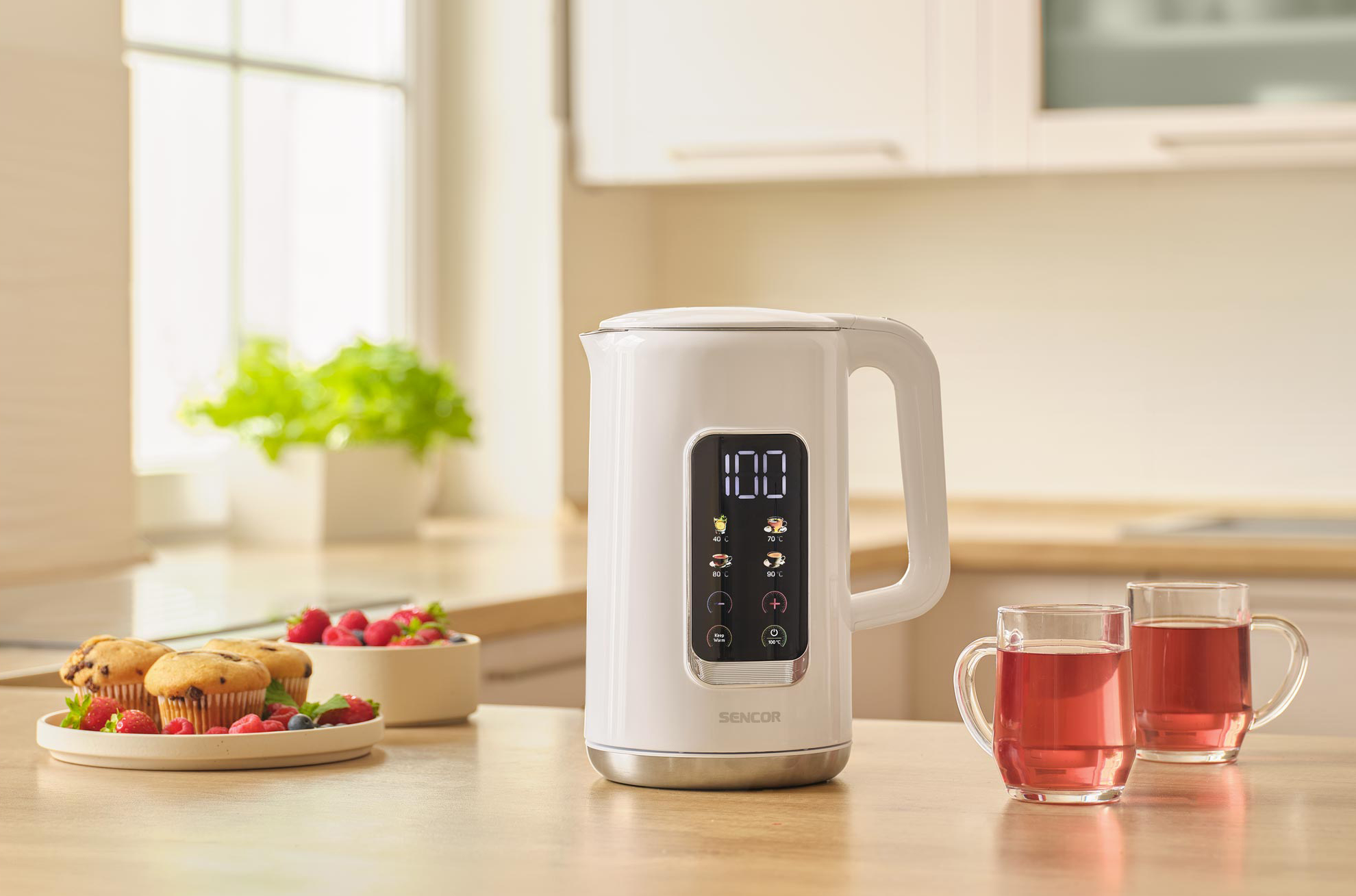 Fehér elektromos vízforraló digitális kijelzővel, két csésze tea, muffinok és málna egy faasztalon.