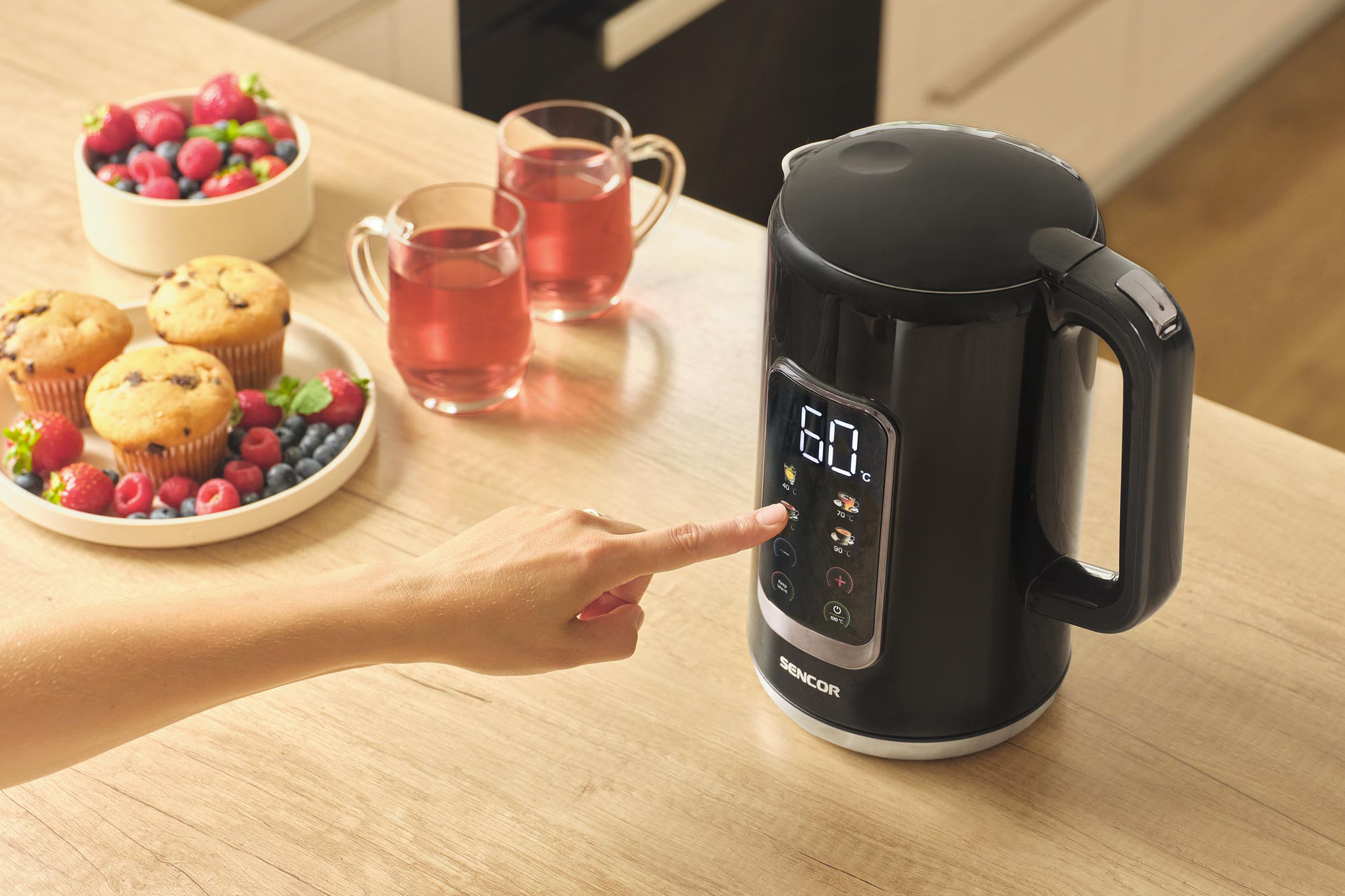 Egy fekete elektromos vízforraló, egy kéz megnyom egy gombot. Bogyók és muffinok.