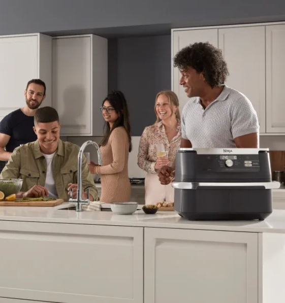 Grupa osób w kuchni. Mężczyzna przygotowuje jedzenie, inni towarzysko spędzają czas.